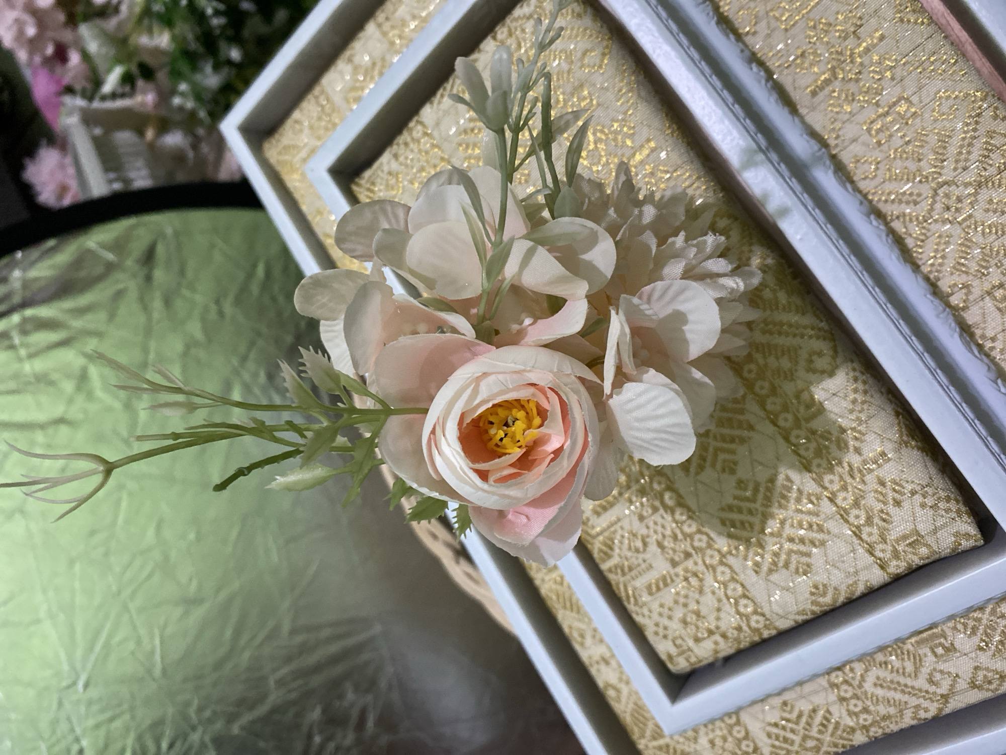 This media file captures a close-up, overhead shot of a decorative floral arrangement placed on a beautifully patterned box. The arrangement primarily consists of artificial flowers in soft pastel shades of pink and white, with delicate green foliage. The main flower in the foreground is a layered bloom with light pink edges and a striking yellow and orange center. Surrounding it are other white flowers with intricate petal details.
The box itself is layered and features a textured, golden fabric with an intricate, traditional pattern, possibly indicating a special occasion such as a wedding or a formal event. The box has white borders that create a framed effect on its surface. The background is softly blurred, with hints of green foliage and other decorative elements, suggesting a setting that is either outdoors or a well-decorated indoor space. To the left of the frame, a reflective, slightly crinkled surface in a muted green hue adds a contrasting texture to the scene.
There is no visible text in the image. The lighting suggests it might be daytime, with soft, diffused light illuminating the subjects without harsh shadows. The overall impression is one of elegance and careful presentation, typical of celebratory decorations. Given the location context of Kuala Pembuang, Indonesia, this arrangement could be part of a local wedding ceremony or a cultural event.
