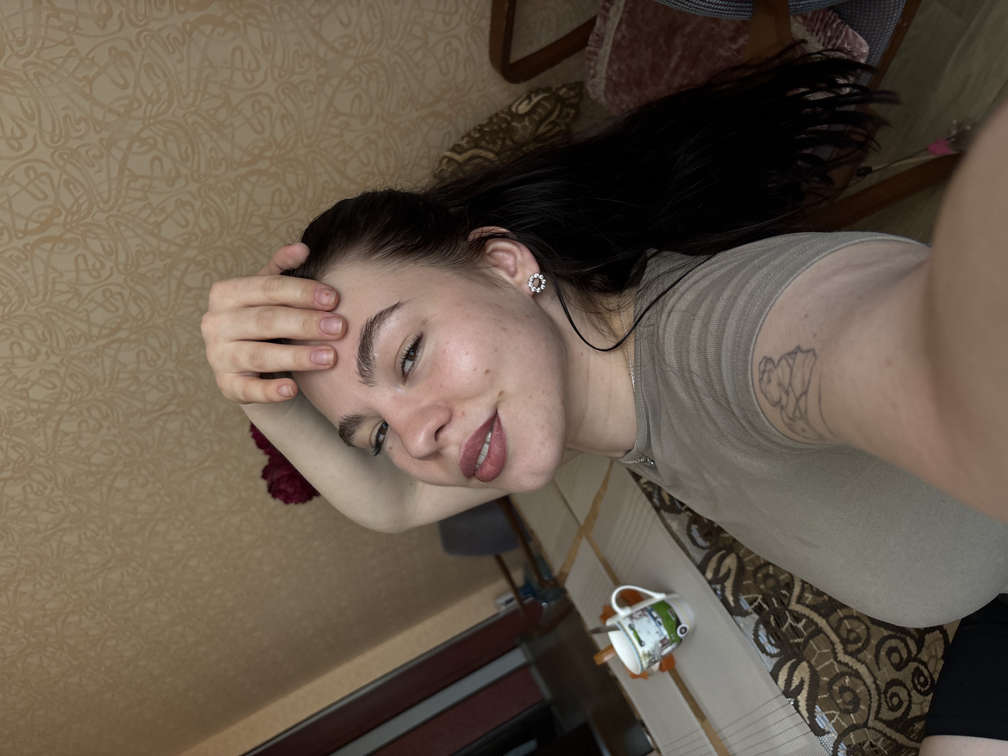 A fair-skinned young woman with dark hair is facing the camera, smiling slightly. She has a hand on her forehead and is wearing a light gray top and a silver necklace. The visible part of her left arm has a tattoo. She is also wearing an earring with a floral design. The background includes patterned wallpaper, a wooden chair, and flooring. A mug with a car design is visible on the floor, alongside a broom. Based on the visible furnishings and style of the room, the location appears to be a domestic interior, possibly in an apartment building. There is no definitive visual information to place the scene specifically in Perm, Russia. The description is limited to the elements visible in the image.