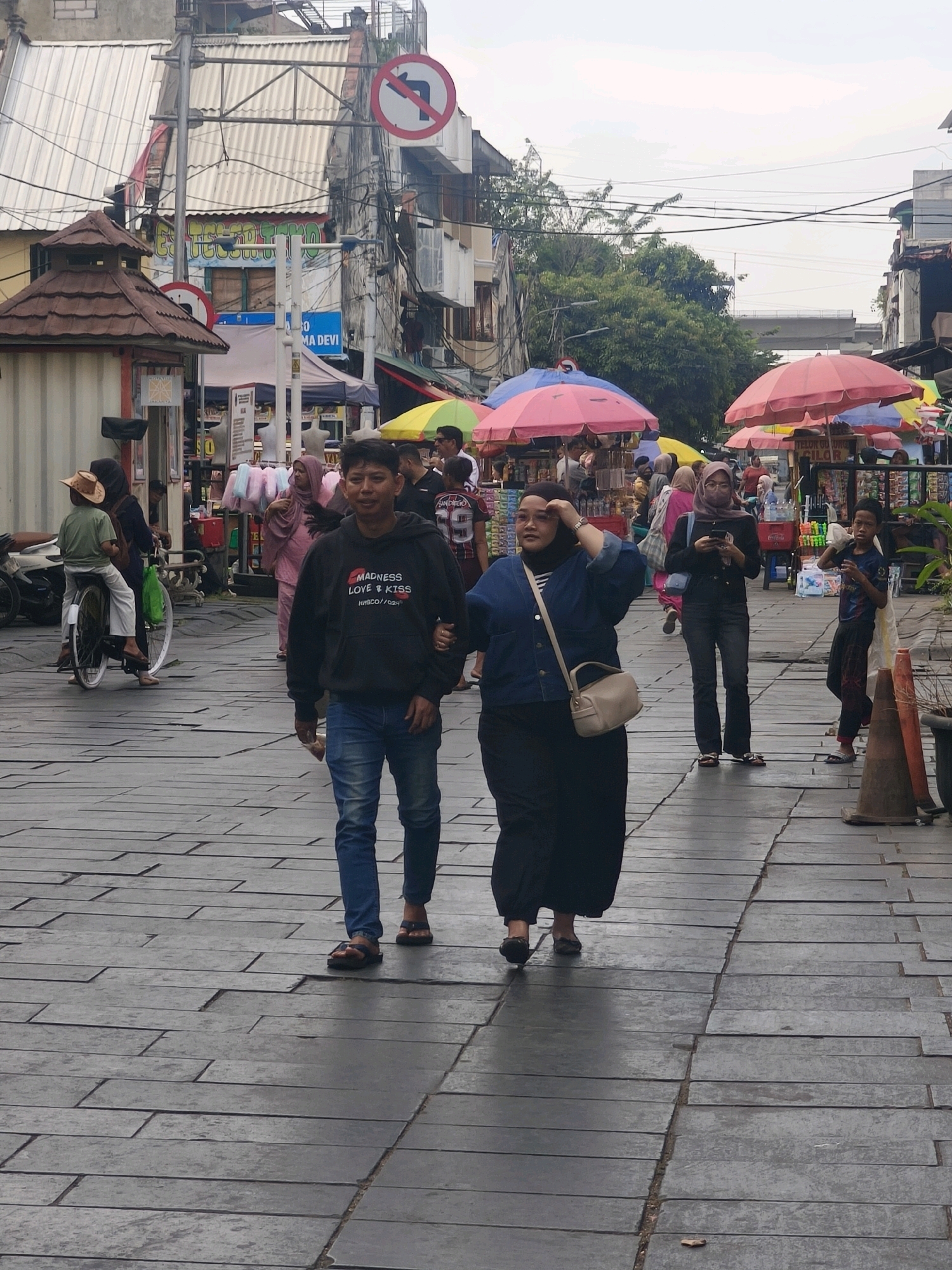 The image depicts a bustling street scene in West Jakarta, Indonesia, likely a marketplace or pedestrian zone. In the foreground, a young man wearing a black hoodie with text and blue jeans walks arm-in-arm with a woman in a dark blue denim jacket and a long black skirt. They are walking on a cobblestone-like paved walkway. Behind them, other people are visible, including a woman in a pink hijab and a woman with a pink hijab and a face mask. Several market stalls with colorful umbrellas are set up, selling various goods, with some displaying mannequins and food items. A man on a bicycle with a green bag attached is seen on the left side of the image. Overhead, electrical wires crisscross, and a no-left-turn traffic sign is visible. The architecture of the buildings lining the street appears to be a mix of older and modern structures. The overall atmosphere is one of lively commerce and pedestrian activity.