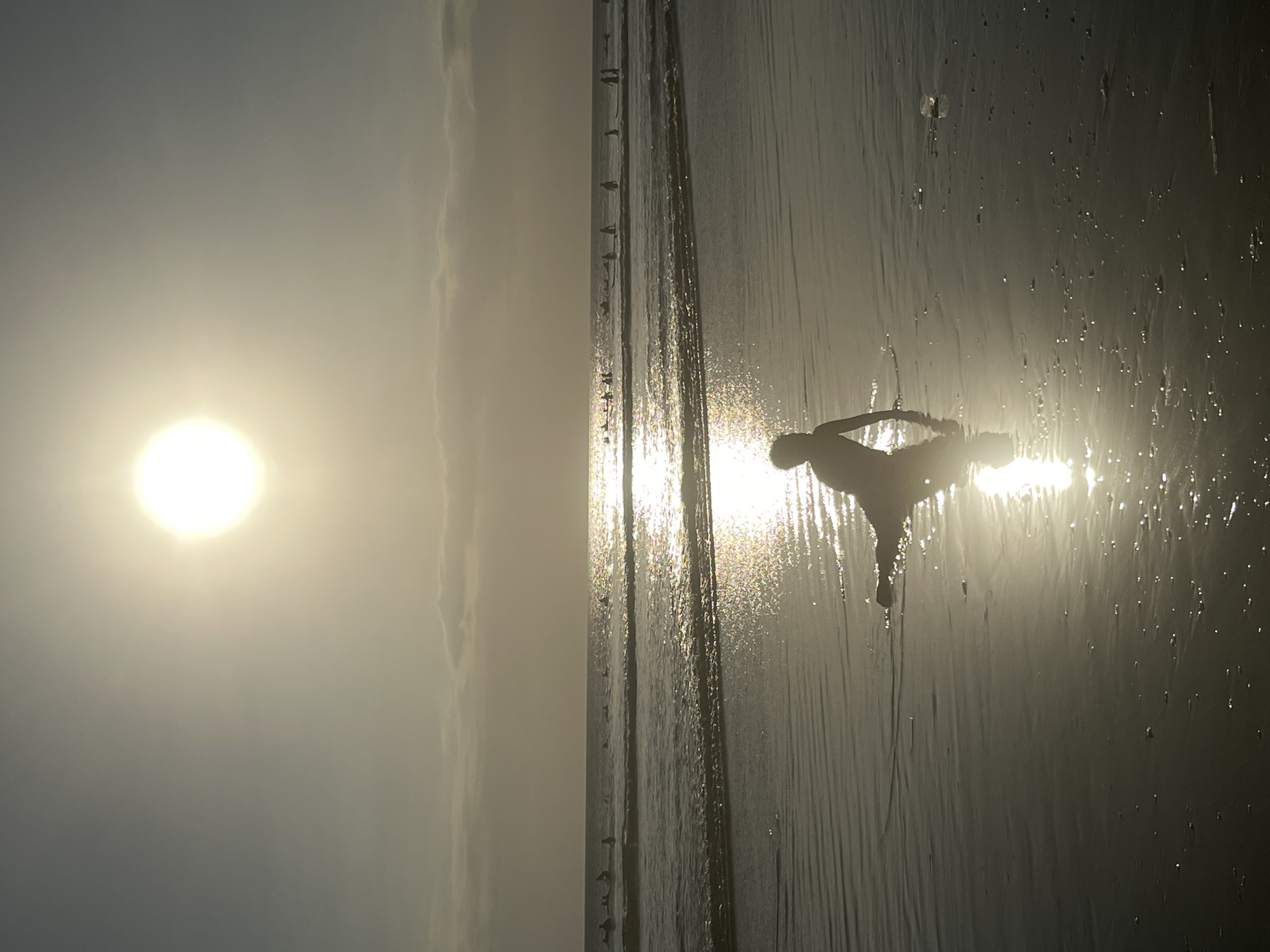 The image shows a silhouette of a person in a yoga pose on a wet sandy beach during sunset. The sun is low on the horizon, casting a bright, hazy light and creating reflections on the water's surface. The person is positioned on the right side of the frame, with their body angled towards the setting sun. The beach appears wet, possibly from receding waves, with small water droplets scattered across the sand. In the distance, a line of small figures, likely birds, are perched along what appears to be a breakwater or pier. The sky is a soft gradient of hazy yellow and grey, suggesting a cloudy or misty sunset. The overall mood is serene and contemplative, evoking a sense of peace and connection with nature. The location is Kuta, Indonesia, known for its beaches and sunsets. There is no visible text in the image.
