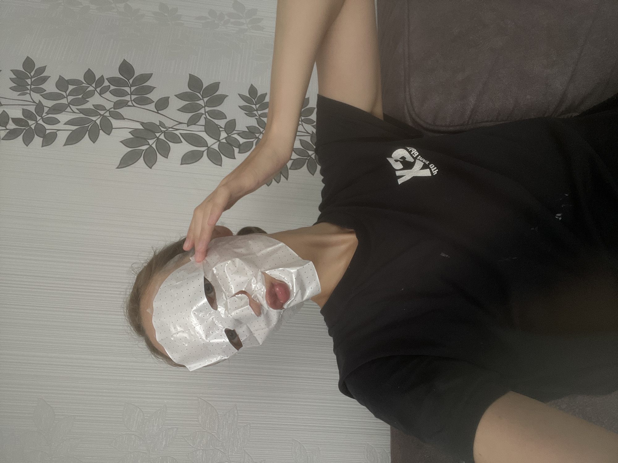 A young woman wearing a black t-shirt and a white sheet mask that covers her face except for her eyes and mouth is sitting on a dark brown couch. Her right hand is raised to her forehead, seemingly adjusting the mask. The background features a light gray, textured wallpaper with a repeating pattern of stylized gray leaves on a vine. The lighting is dim, and the scene appears to be indoors, likely a home setting. The image does not contain any specific landmarks or indicators that would suggest its location within the city of Sharkan, Russia.