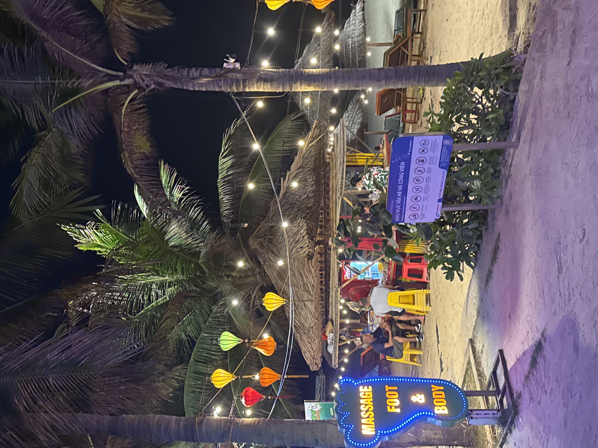 This is an image of a beachside scene in Đà Nẵng, Vietnam, at night. A "MASSAGE FOOT & BODY" sign with blue lights is on the right. Palm trees line the foreground and left of the frame. Colored lanterns are strung between the palm trees, and lights illuminate a thatched-roof structure in the center, likely a restaurant or bar. People sit at colorful tables and chairs beneath the thatched roof. A sign with Vietnamese text and symbols is visible to the right. The ground is sand, suggesting a beach location.