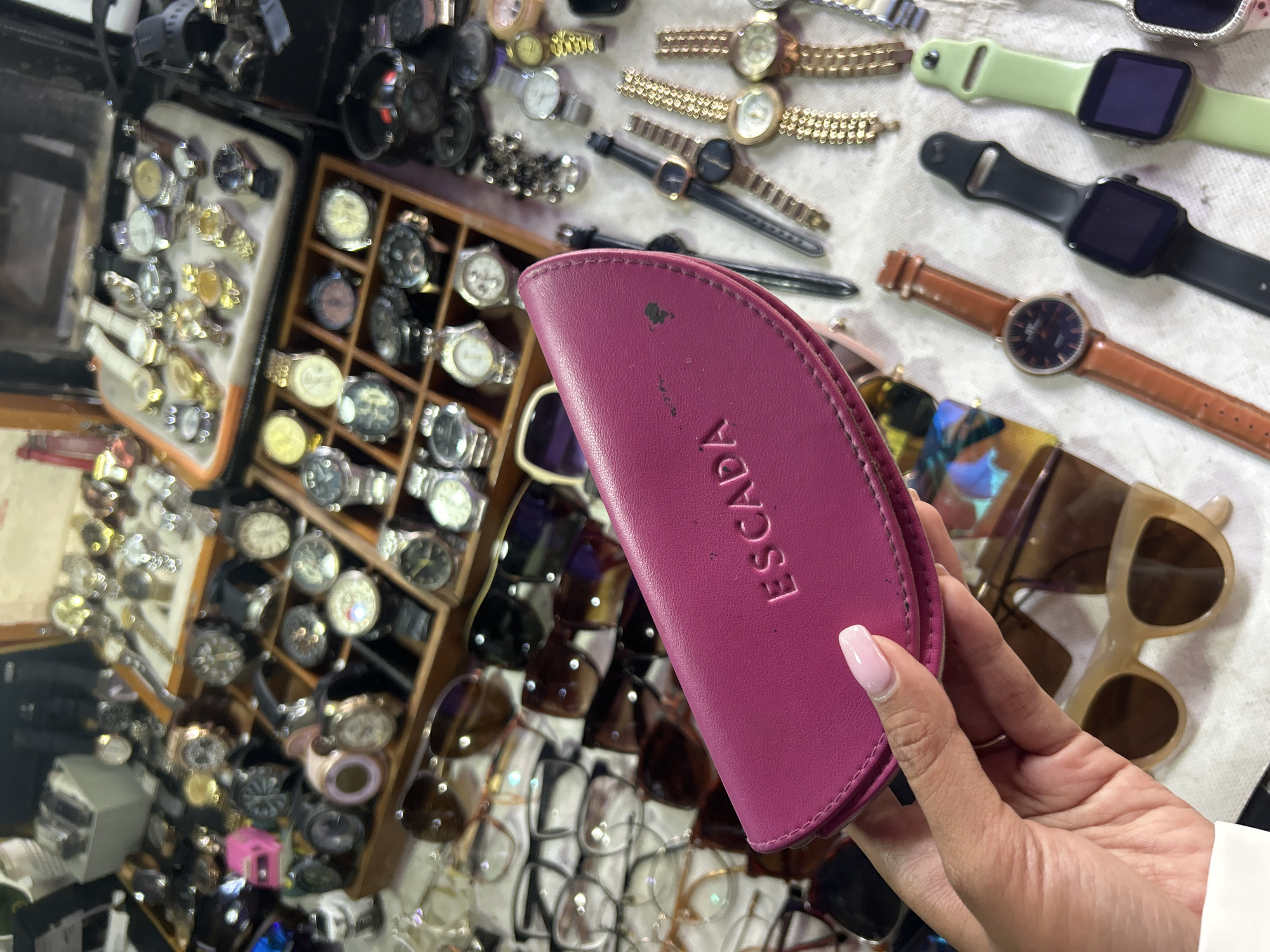 In Denpasar, Indonesia, a hand with a light pink manicure holds a magenta-colored case, possibly for glasses, with the embossed word "ESCADA" visible on it. The case is positioned in the foreground of the image. Behind the hand and case, a collection of various wristwatches is displayed on a white surface. Several watches are arranged in rows, featuring different band materials like leather and metal, and watch faces of varying designs and colors. Some watches are gold-toned, while others have darker bands. In the background, more watches are visible in wooden display boxes. Additionally, several pairs of eyeglasses, with both clear and tinted lenses, are scattered amongst the watches, suggesting a retail or market setting where accessories are sold.