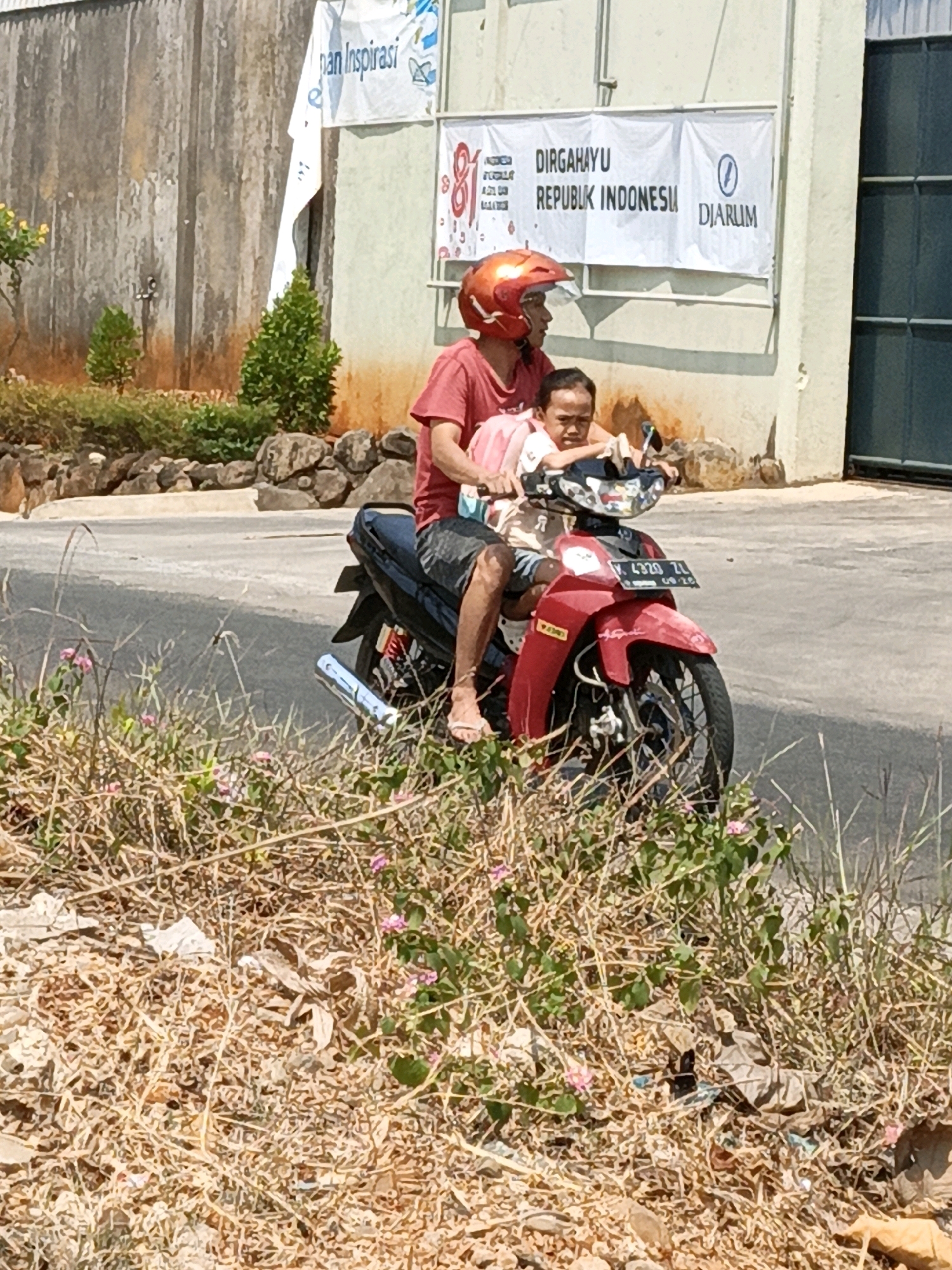 A man wearing a red t-shirt and an orange helmet rides a red motorcycle. He is carrying a child, who is wearing a pink shirt and sitting in front of him. Both individuals appear to be looking forward. The motorcycle has a license plate that reads "4320 ZL". They are riding on a paved road next to a grassy area with some pink flowers. In the background, there is a wall with a banner that says "DIRGAHAYU REPUBLIK INDONESIA". This scene is likely located in Kuwasen, Indonesia, given the presence of Indonesian text on the banner. The environment appears to be outdoors on a sunny day.