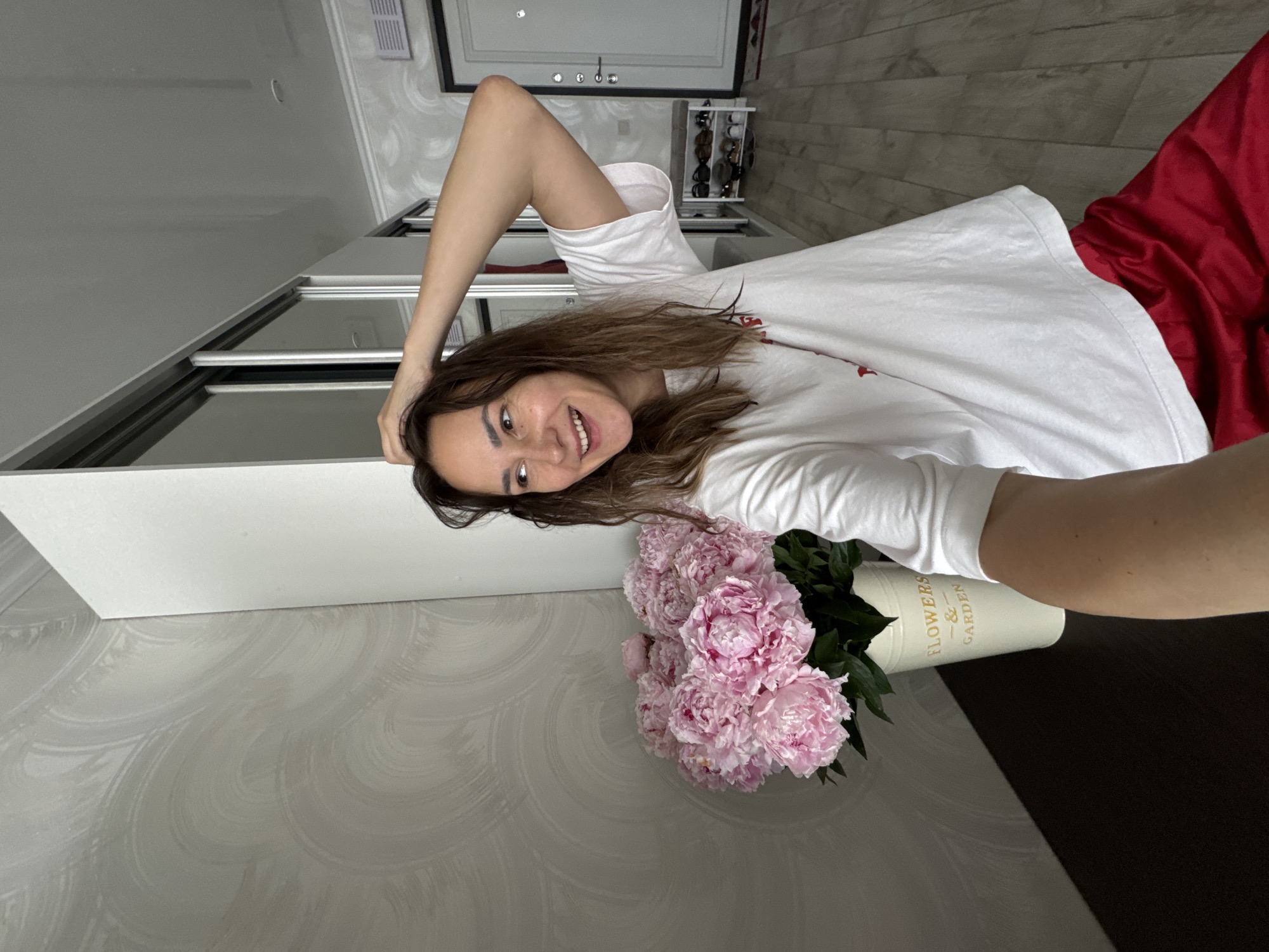 A woman with brown hair is standing in a hallway, taking a selfie. She is wearing a white t-shirt and red pants. She has her right hand on her head, and she is smiling. To her left, there is a bouquet of pink peonies in a white pot with "FLOWERS & GARDEN" written on it. The hallway has patterned wallpaper and a large white wardrobe with mirrored doors. There is a shoe rack with shoes in the background, and a white door. The floor is wooden. The scene is indoors and appears to be in a residential setting.
