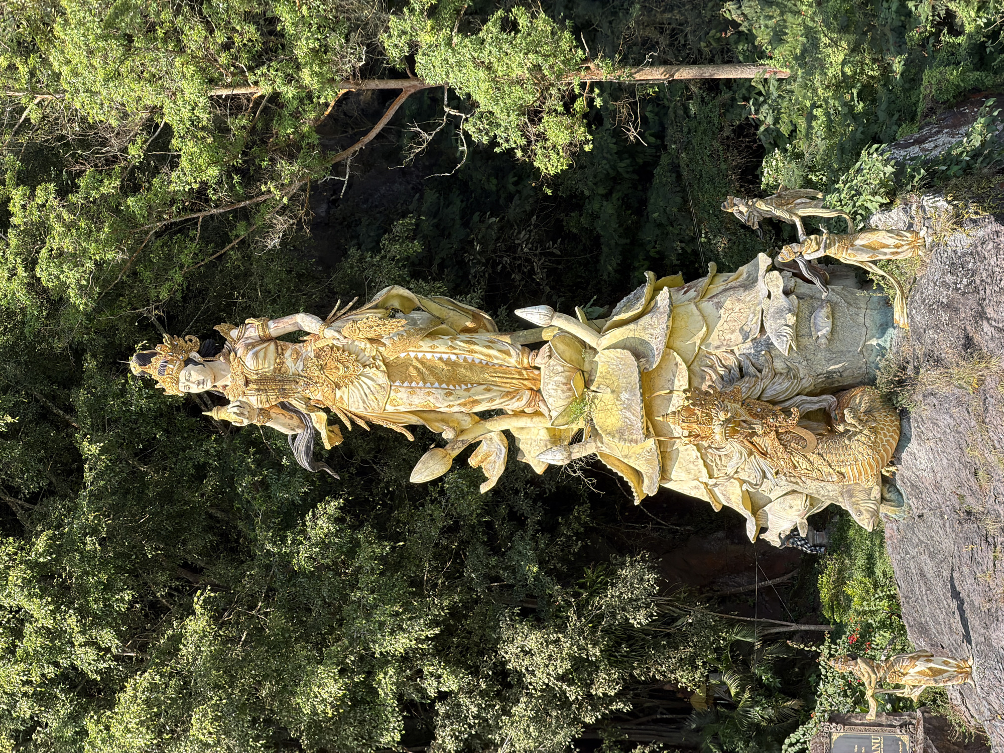 The image shows a large, ornate golden statue of a woman, adorned in a crown and traditional attire, standing on a layered, petal-like base. To the lower left of the main statue, a smaller golden statue of a woman stands on a rock, holding a staff. To the lower right of the main statue, two more smaller golden statues of women are positioned on a rocky outcrop. At the base of the main statue, a golden dragon sculpture is coiled around a layered structure, with fish depicted on its side. The statues are situated on a rocky slope with lush green trees and foliage forming the background. A sign with text is partially visible on the left side of the image. The scene appears to be outdoors, under natural lighting, with dappled sunlight filtering through the trees. This statue is located in Kintamani, Bali, Indonesia, specifically at the Brahma Tirta Sari Temple.