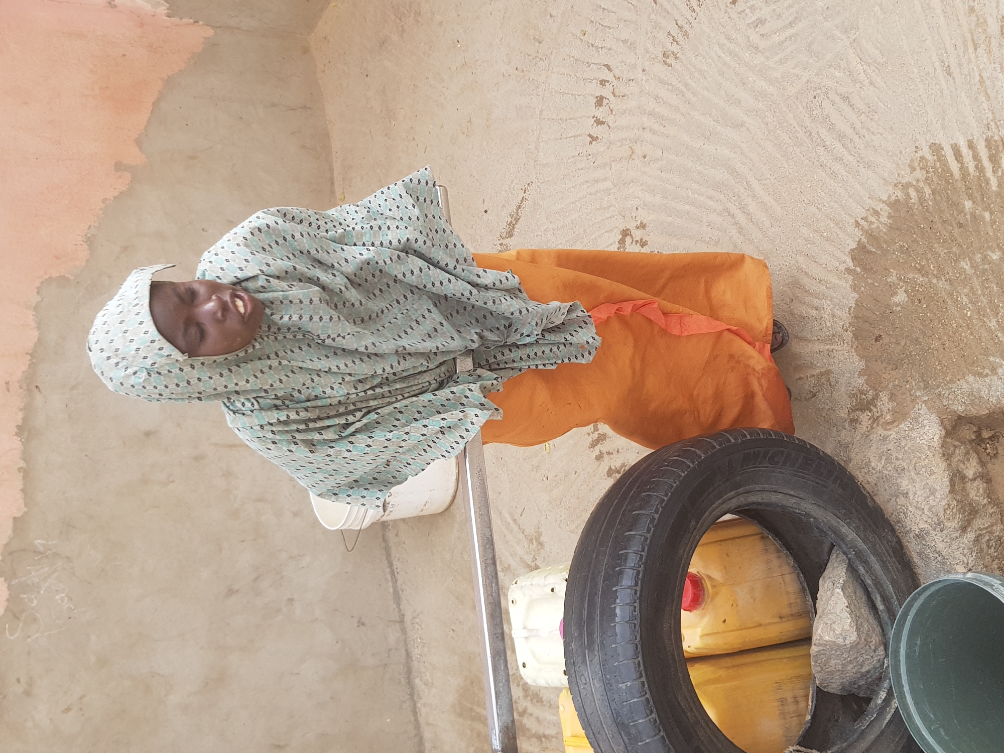 An image of a woman, likely from the city of Dutsinma A, Nigeria, wearing a patterned headscarf and an orange dress. She is seated on a white plastic chair. A tire labeled "MICHELIN" leans against containers. A green bucket is present, and the setting appears to be an outdoor area with a textured wall.