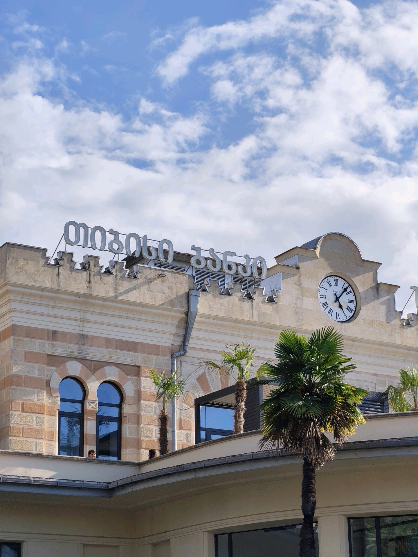 This is a medium-angle, vertical outdoor shot of the Batumi train station in Batumi, Georgia. The building has a light-colored facade with alternating bands of light pink and beige stone. Above the windows, there are decorative crenellations along the roofline. A large clock with Roman numerals is mounted on the facade, indicating a time of approximately 3:10. Large, illuminated Georgian letters spell out "ბათუმი" (Batumi) on the roof. Several palm trees are visible in front of and on the terraces of the building. The sky above is blue with scattered white clouds. Two people are visible through one of the arched windows.