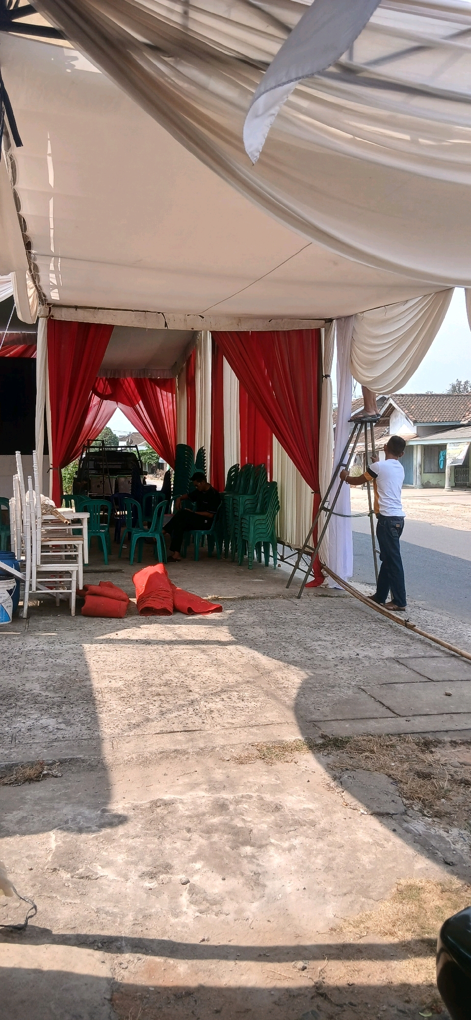 The image captures an outdoor scene in Banjar Rejo, Indonesia, under a large tent. The tent's interior is draped with alternating red and white fabric. Green plastic chairs are stacked in piles, with some spread out and occupied by two individuals. One person, wearing a dark shirt and pants, is seated and appears to be resting or waiting. Another person, dressed in a white t-shirt and blue jeans, stands on a ladder, seemingly engaged in setting up or adjusting the tent's fabric. Rolled-up red mats lie on the concrete ground in the foreground. A vehicle, likely a truck, is visible through an opening in the tent. The environment suggests preparation or setup for an event, with the sun casting shadows on the concrete. The background shows houses and a street.