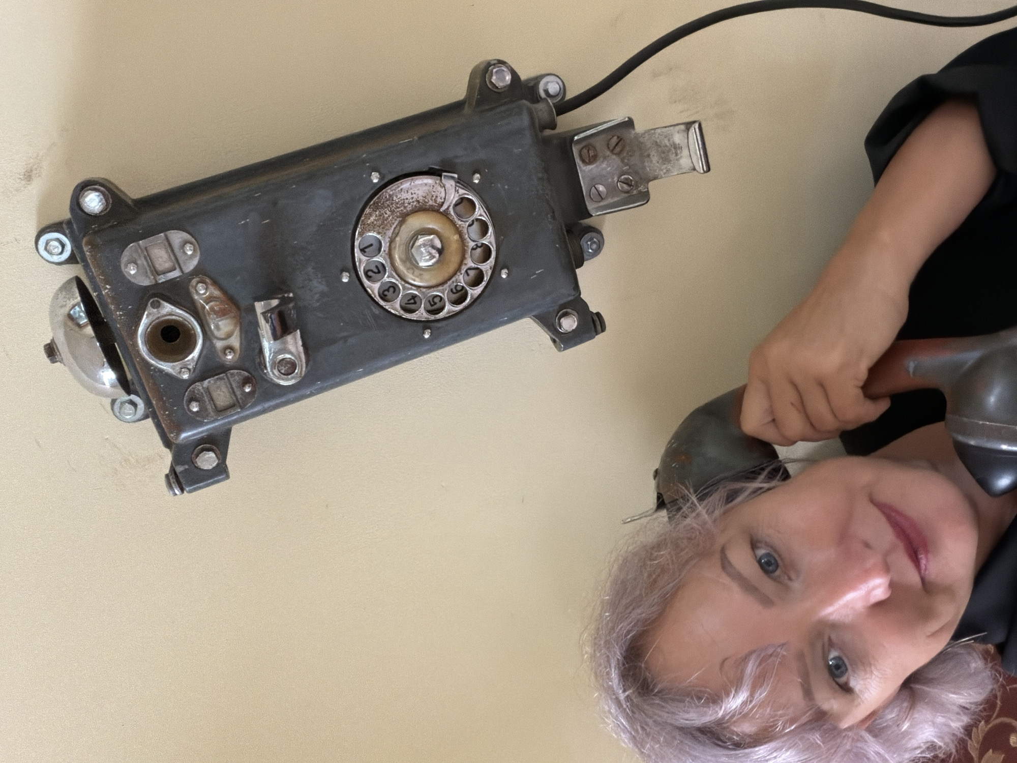 A medium shot captures a woman with lavender-colored hair holding a vintage rotary telephone receiver to her ear. The woman is positioned in the lower left quadrant of the frame, her face turned towards the camera. She is wearing a black garment. To her right, mounted on a light beige wall, is an antique, dark gray wall-mounted telephone. The telephone features a prominent rotary dial with numbers, a metal bell at the top, and various metallic components and screws. A black cord extends from the telephone downwards and out of frame to the right. The background consists of the plain beige wall. The scene does not contain any identifiable elements that place it specifically in Sochi, Russia.
