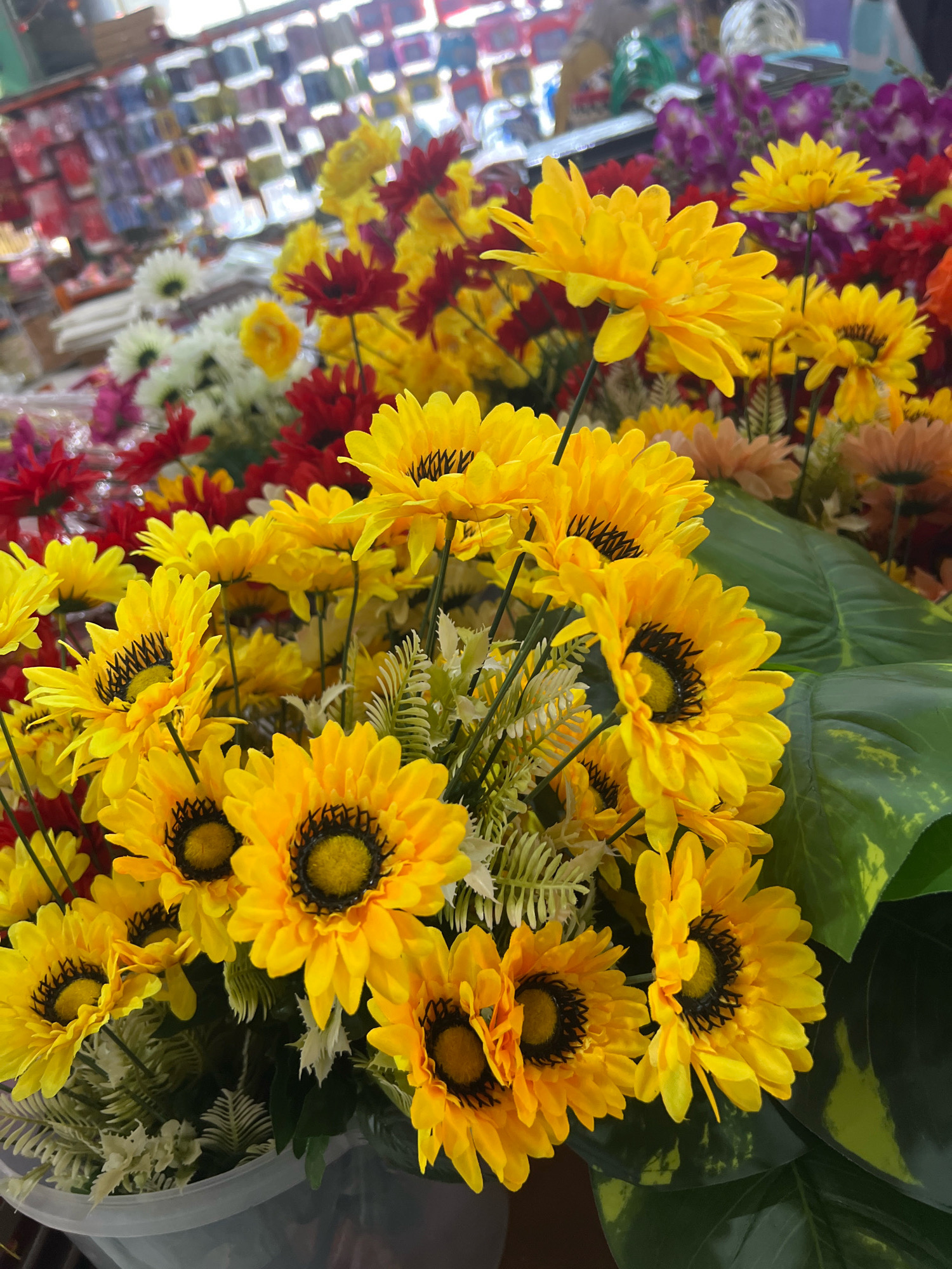 A close-up, eye-level shot captures a vibrant display of artificial sunflowers and other flowers. The sunflowers, predominantly yellow with black centers, are clustered together, filling the foreground and midground. Their petals are slightly ruffled, and some have a subtle sheen. Interspersed among them are red and white flowers, as well as hints of purple flowers in the background, creating a colorful and textured arrangement. Large, dark green leaves with lighter green veins are visible on the right side, adding depth to the composition. The flowers appear to be arranged in a pale grey bucket, partially visible at the bottom of the frame. In the blurred background, shelves are stocked with rows of small, rectangular items in various colors, suggesting a retail environment like a flower shop or craft store. The overall lighting is bright, possibly from overhead lights or natural light filtering in from a window, creating soft highlights on the petals. The scene is a still life, devoid of any human presence or action. This scene is likely located in a flower shop in Denpasar, Indonesia, given the common availability of such artificial flower arrangements in local markets and stores across the city.