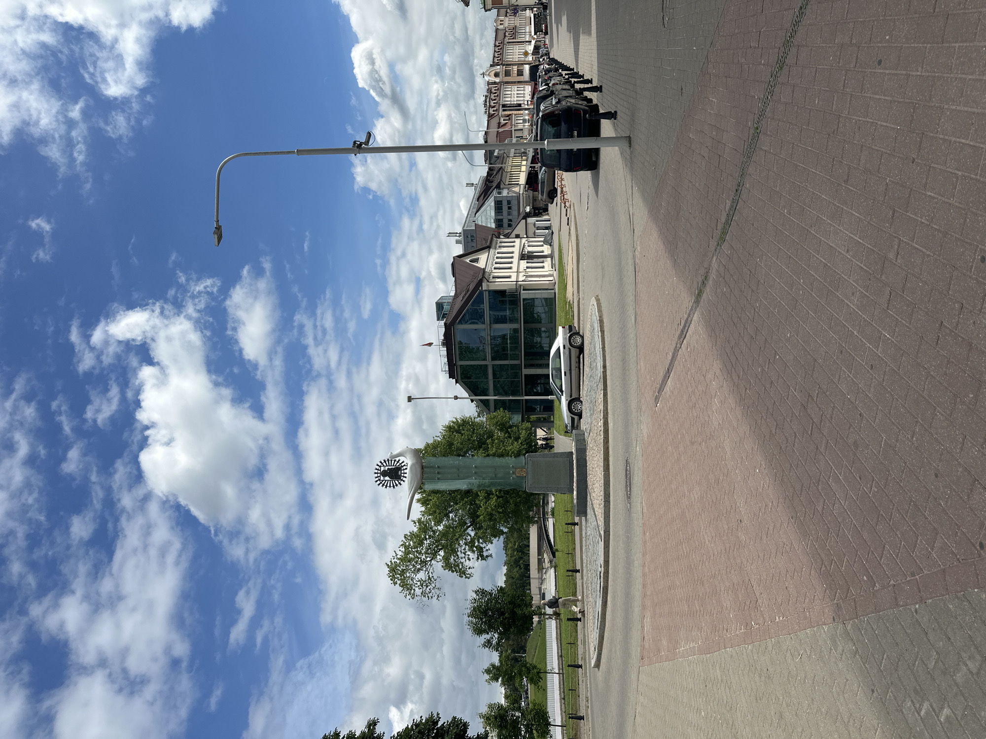 This image depicts a street scene in Minsk, Belarus, likely in a public square or intersection. The foreground is dominated by a paved roundabout with red and grey brickwork. A modern-looking sculpture of a bird with a sunburst design on a tall, translucent green pedestal stands prominently in the center of the roundabout.
To the left of the roundabout, a lone figure, possibly a man wearing light-colored trousers and a dark shirt, walks away from the camera. Behind him, trees and a body of water or a canal can be seen, with a bridge in the distance.
On the right side of the image, a street lined with parked cars runs parallel to the sidewalk. The cars are varied, including black vans and a silver sedan. The sidewalk is also paved with brickwork. Beyond the parked cars, there are several buildings, characterized by their architectural styles, some appearing older with multi-story facades, while others have more contemporary glass and modern designs. A street lamp with a camera attached is visible on the right side of the street, positioned near the lamp post.
The sky is blue with scattered white clouds, indicating fair weather. The overall impression is of an urban environment with a mix of historical and modern architecture, showcasing a public space with a notable monument.