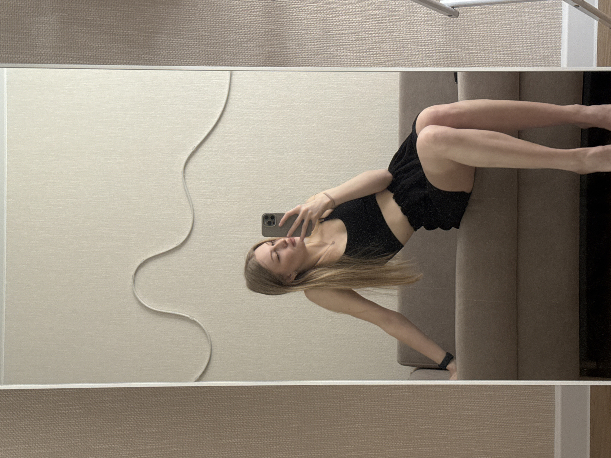 A young woman is reflected in a rectangular mirror. She holds a smartphone up to take a selfie. The woman has long blonde hair and is wearing a black crop top and shorts. She is sitting on a beige couch. A white cord is visible on the wall behind her. The mirror is framed with white trim. The top and bottom of the mirror reflect the beige textured wall of the room. There is no visual information in the image to verify that the location is Perm, Russia.