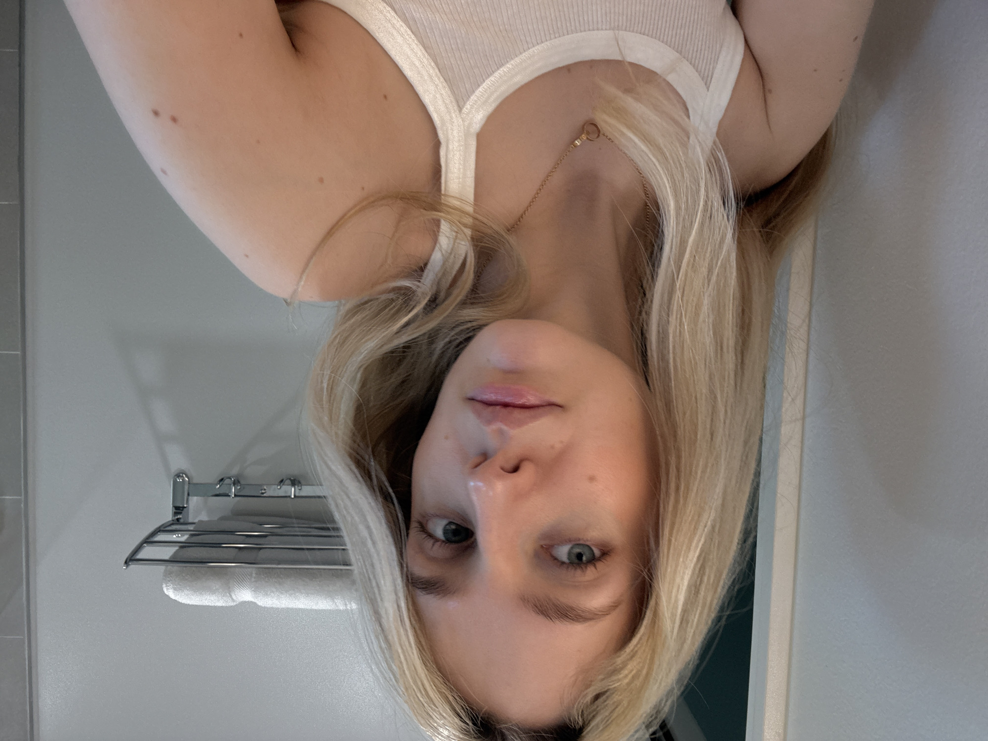 A young woman with blonde hair and blue eyes is captured in a close-up, self-taken photograph. She is wearing a white ribbed tank top and a delicate gold necklace with a circular pendant. Her skin is fair, with a few moles visible on her right arm and shoulder. The background appears to be a bathroom, with a wall-mounted chrome towel rack holding a neatly folded white towel. A portion of a gray tiled wall is also visible to the right. The lighting is soft and natural. The image does not provide any specific indicators of its location within Miami, United States of America.