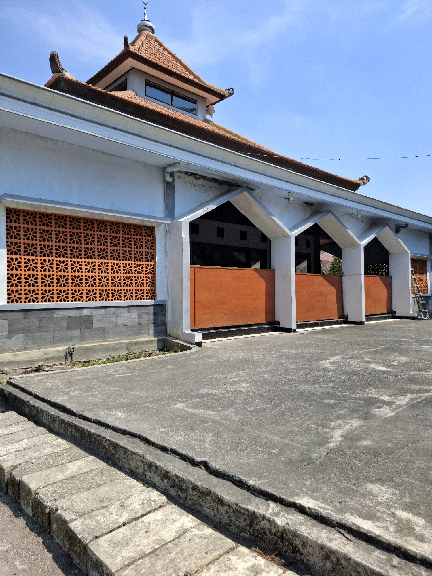 The image shows a mosque in Kedungmaron, Indonesia. The mosque has a light blue facade with orange decorative vents. The roof is tiled with terracotta tiles and features a traditional Indonesian architectural style. The foreground shows a paved courtyard with concrete borders. The sky is clear and blue. There are no people visible in the image. The scene appears to be well-lit by natural sunlight.