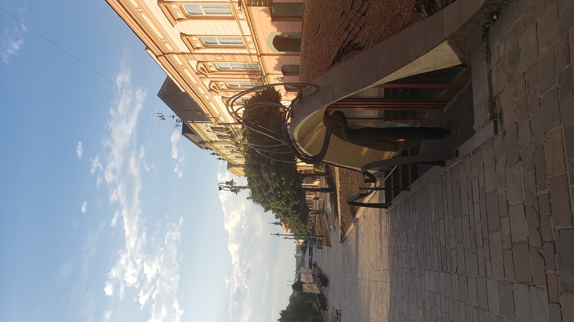 The image captures a view down a paved pedestrian walkway lined with trees and buildings on the right, likely in the historic city center of Prešov, Slovakia. The walkway is made of irregular stone pavers and features several benches and lampposts. On the right side, a row of light-colored, ornate buildings with arched doorways and windows can be seen. In the foreground, a unique, modern-looking structure with a curved, light-colored roof and dark metal railings leads down to an entrance. The sky is blue with scattered white clouds, and the sun is casting long shadows, suggesting it is late afternoon or early morning. There are no people clearly visible in the scene, but the presence of benches and outdoor seating areas in the distance implies a public space.