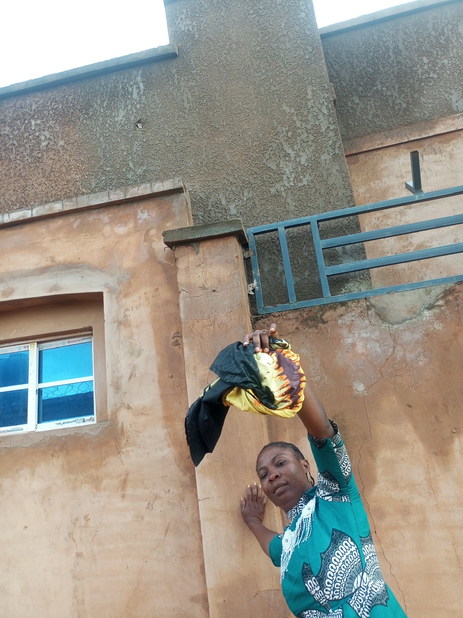 A medium shot captures a woman in Tempure, Nigeria, leaning against a textured, tan-colored building. The woman, with dark hair braided, wears a green dress with a white and black patterned design. She holds a colorful piece of fabric, possibly a headscarf or wrap, in her raised right hand. The building features a white-framed window with blue glass and metal bars. A dark metal railing runs horizontally along the upper portion of the building. The sky is visible in the top left corner, appearing bright white. The scene is shot from a low angle, looking up at the woman and the building.