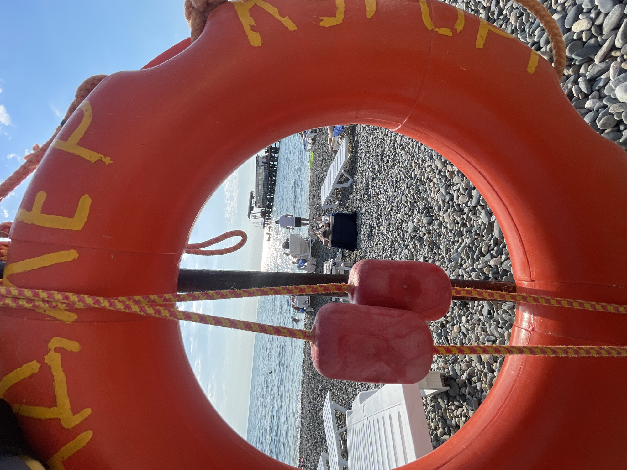 An extreme close-up shot features a bright orange life preserver in the foreground, with yellow lettering partially visible. Two red buoys hang from yellow and red striped ropes attached to the life preserver. The background reveals a pebbled beach, the sea, and a pier in the distance, bathed in sunlight. Several people are visible on the beach, some reclining on lounge chairs, while others are near the water's edge. The scene appears to be a sunny day at a beach resort in Sochi, Russia, likely in the Adler district given the presence of a pier and beach amenities characteristic of the area.