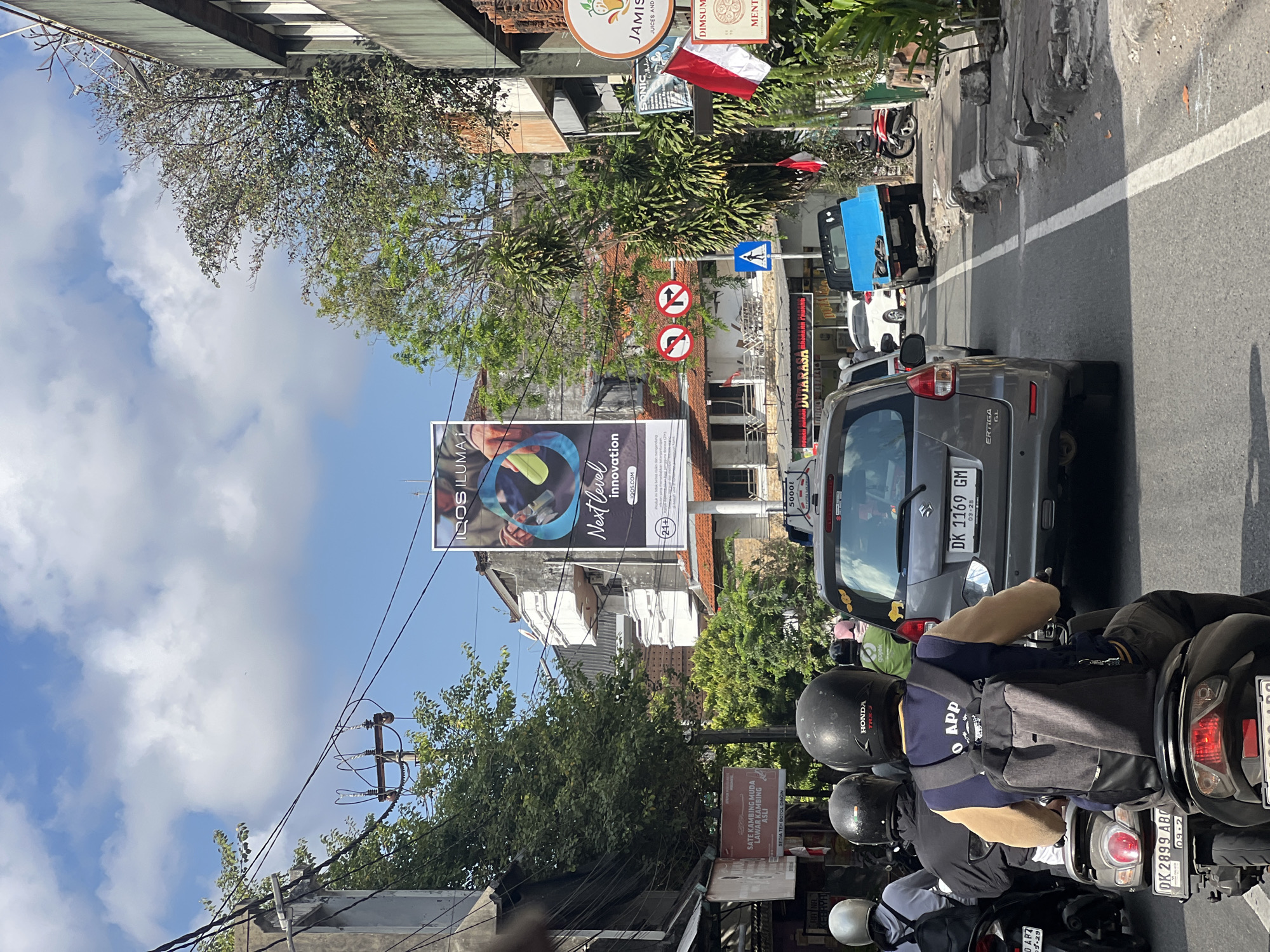 This is a photograph taken from a moving vehicle on a street in Denpasar, Indonesia, likely during the daytime given the bright sunlight and clear sky with scattered clouds. The street is congested with traffic, predominantly motorbikes, and some cars.
In the foreground, a person on a motorbike is visible from behind, wearing a black helmet and a blue and beige jacket with a backpack. Another person on a motorbike with a white helmet is also in the frame. Several other motorbikes and a grey car are ahead, indicating moderate traffic flow.
Buildings line both sides of the street. On the left, there are low-rise structures with signs, including one with Indonesian text that reads "SATE KAMBING MUDA LAWAK KAMBING ASLI". There are also overhead electrical wires and poles. On the right, a larger building features an advertisement for "IQOS ILUMA" with the tagline "Next Level Innovation". Further down the street on the right, there are more shops and restaurants, with signs for "JAMIS JUICES AND REFRESHMENTS" and "MINT". Indonesian flags are also visible.
The road surface appears to be asphalt, with white lane markings. Traffic signs are present, including directional signs and a pedestrian crossing sign. A bright blue pickup truck is parked on the side of the road, and a red motorbike is also visible. The general environment suggests a bustling urban street scene typical of a Southeast Asian city.
