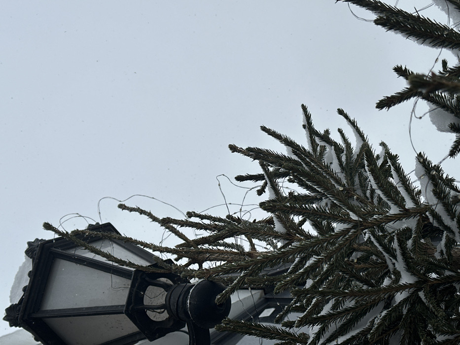 An upward-angled view captures an ornate black wall-mounted streetlamp and evergreen tree branches during a winter snowfall in Kazan, Russia. The cast-iron-style streetlamp, positioned in the upper left, features a hexagonal lantern housing with white or frosted glass panels, topped with a layer of snow. Behind and to the left of the lamp, a building's white-trimmed roofline is visible, also with snow accumulation. Below and to the right of the lamp, the dark green needles of an evergreen tree are partially covered with fresh white snow. Thin, bare wires, possibly remnants of decorative lighting, are intertwined through the tree branches and draped around the lamp housing. The background consists of a uniformly light gray, overcast sky, with numerous small, blurred white specks indicating falling snowflakes. No people or overt actions are depicted.