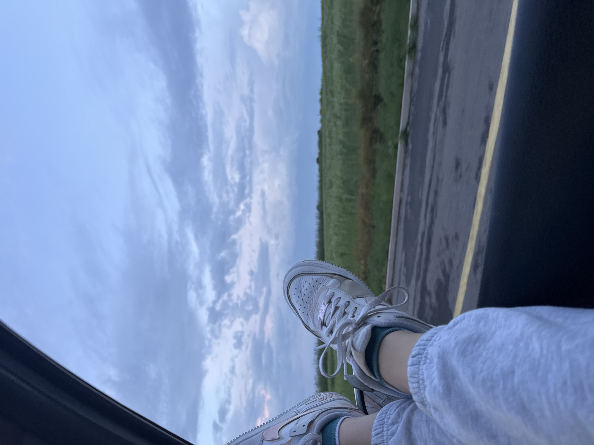 A person with their feet propped up on a vehicle window, facing outward, is depicted in the image. The individual is wearing white sneakers, teal socks, and light gray sweatpants. Outside the vehicle, there is a blurred view of a road with a yellow line marking, a green field, and a cloudy sky. The image does not contain any landmarks that are definitively within the city of Burgas, Bulgaria, so I cannot confirm the location.