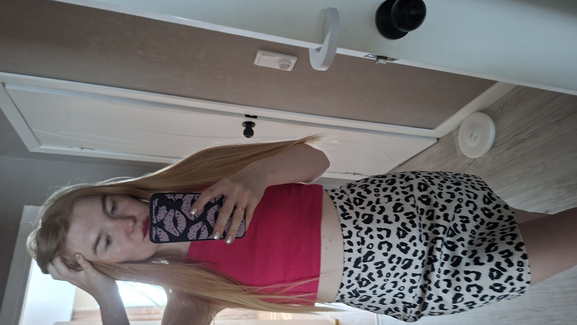 A young woman with long, light-brown hair is taking a selfie in what appears to be a bathroom. She is wearing a red top and a leopard-print skirt. Her phone case has a lip design on it. The bathroom has a white door with a black doorknob, a white baseboard, and light-colored flooring. The reflection suggests this is a mirror selfie. The location is Chaikovsky, Russia.