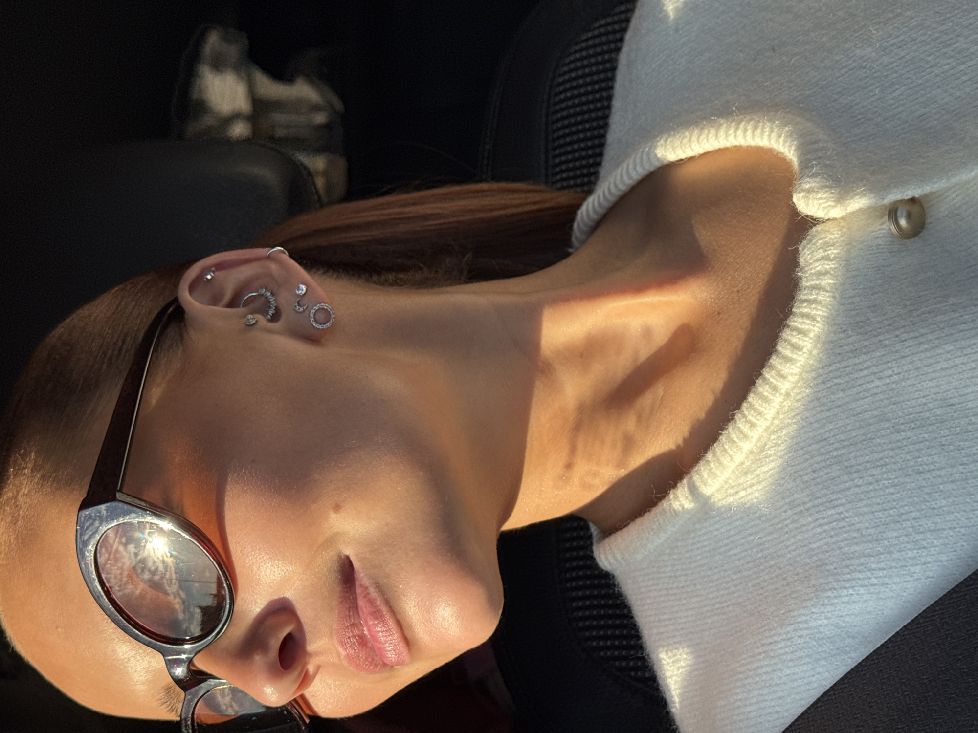 A medium shot captures a woman's profile as sunlight illuminates her face and neck. She wears dark-rimmed sunglasses with reflective lenses, and several silver earrings adorn her ears, including hoops and studs. Her lips are slightly parted, and her skin has a warm, sun-kissed glow. She is wearing a white knitted cardigan with a pearl button. The background is dark and out of focus, suggesting an indoor setting or evening light. There is no discernible connection to the city of Chaikovsky, Russia.