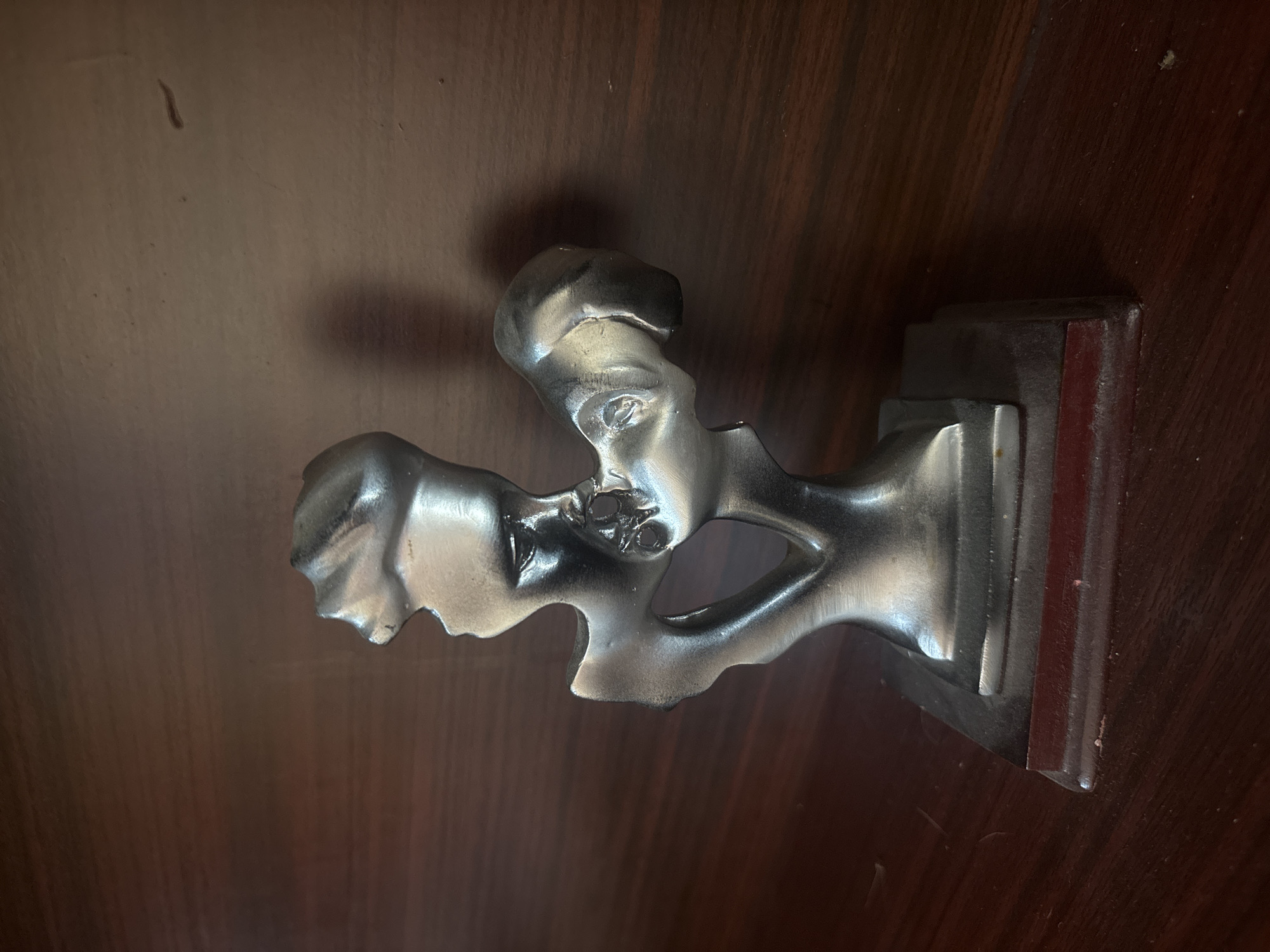 This media file features a metallic, abstract sculpture positioned on a dark wooden surface. The sculpture depicts two stylized human profiles or faces, intertwined and facing opposite directions. One face is more distinctly rendered, facing left, with visible details like an eye and the curve of a nose and forehead. The other face is less defined, seeming to emerge from or merge with the first, facing towards the right. The sculpture has a shiny, silvery finish, possibly of pewter or polished aluminum, which reflects light and highlights its curved and angular forms.
The sculpture is mounted on a dark, rectangular wooden base that features stepped edges. This base rests on a broader, dark brown wooden surface, which exhibits visible grain patterns and a smooth finish. Small dust particles are scattered on this surface, suggesting an indoor setting. The lighting creates subtle shadows, emphasizing the three-dimensional nature of the sculpture.
No activity is taking place; the image is a static portrayal of an art object. There is no visible text. The location context for this item is Asenovgrad, Bulgaria. The overall presentation is artistic, with a strong contrast between the metallic luster of the sculpture and the warm, dark tones of the wood.