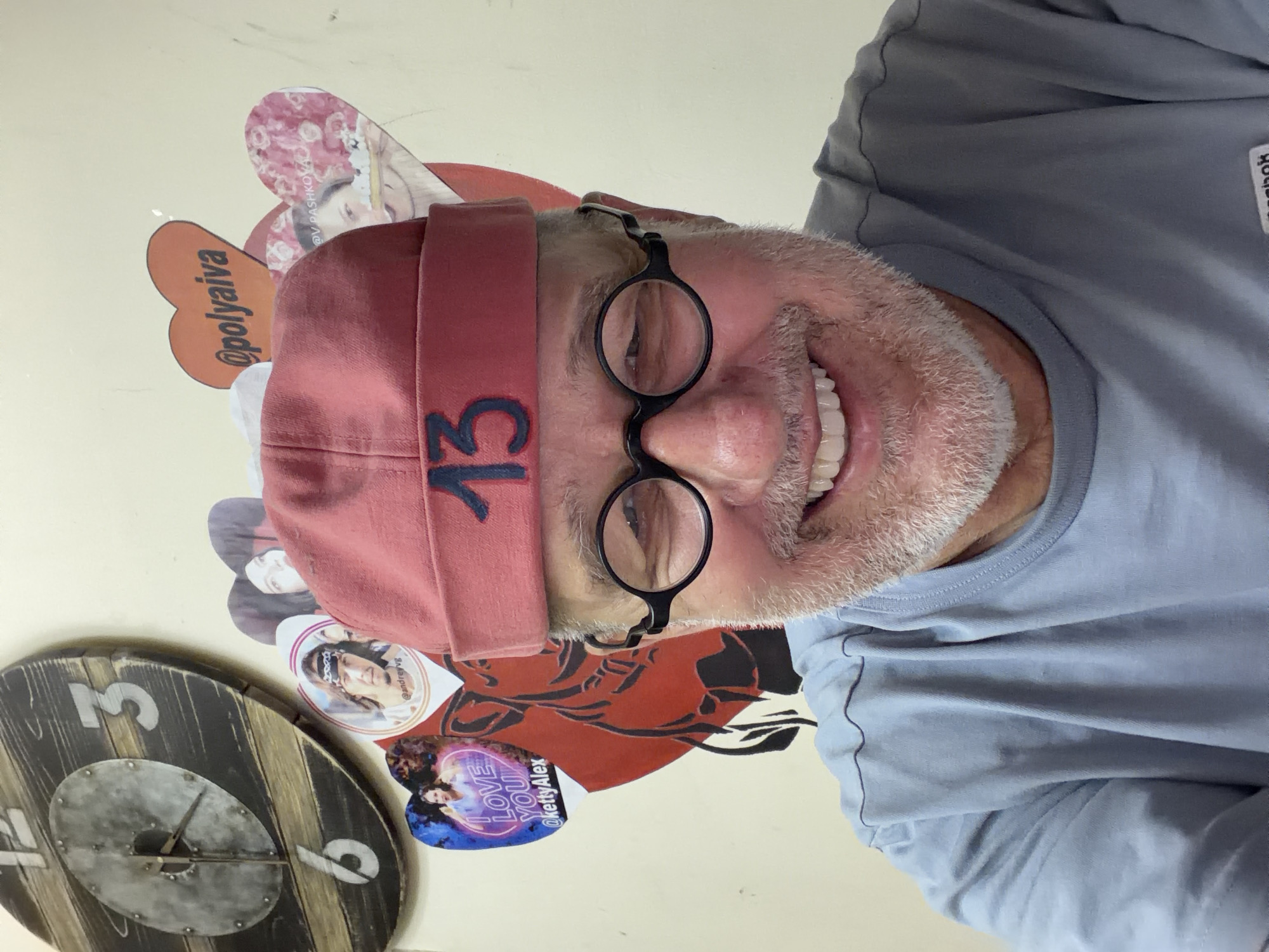 A man with graying facial hair and a red beanie with the number "13" embroidered on it smiles at the camera. He wears round-rimmed glasses and a light blue t-shirt. Behind him, a large wooden clock with a distressed finish hangs on a beige wall. Several heart-shaped stickers are also visible on the wall, some with photos of people and social media handles.