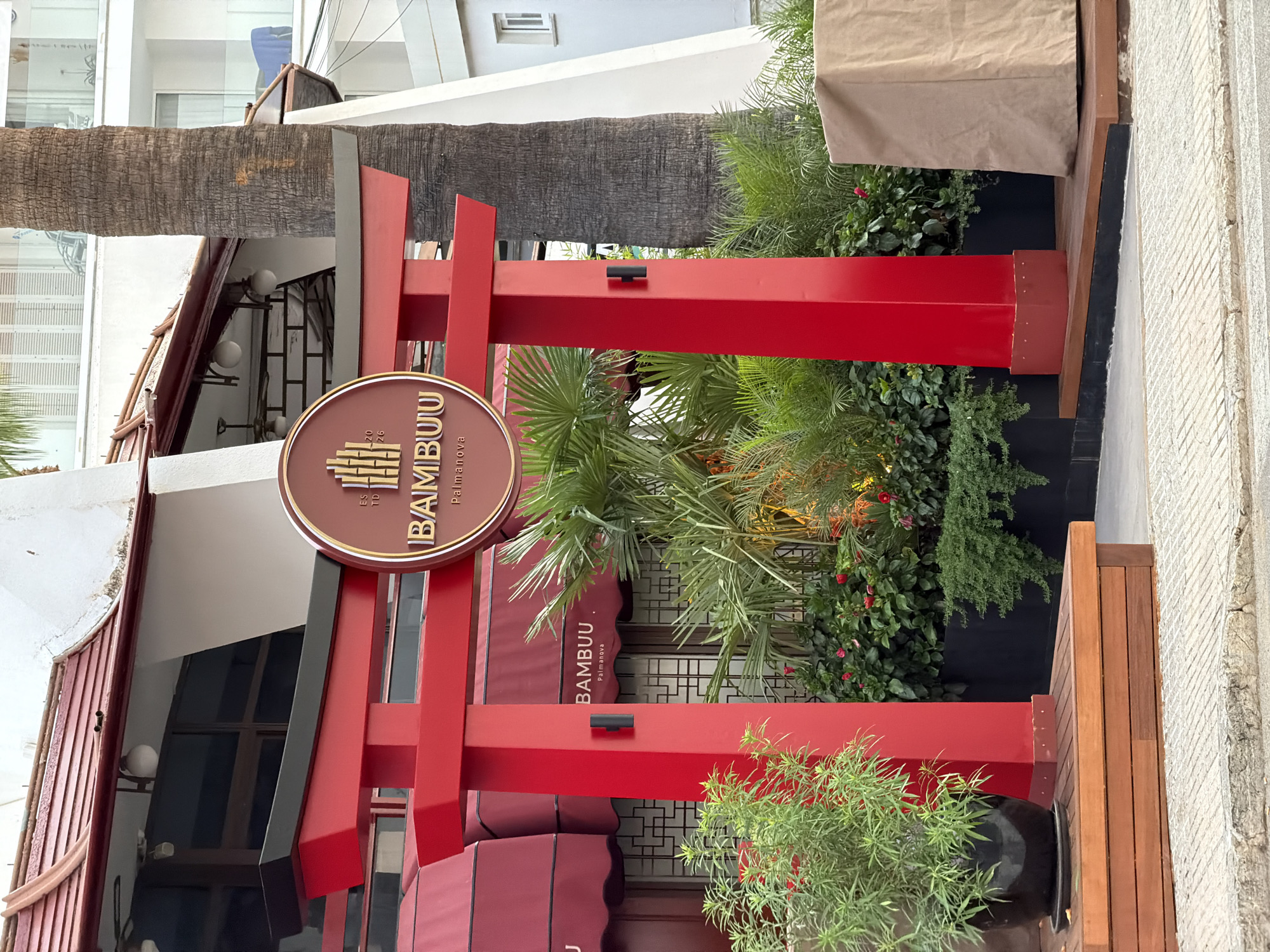 The image shows the exterior of a restaurant called "BAMBUU" in Palmanova, Spain. A vibrant red torii gate, a traditional Japanese gate, stands prominently in the foreground, marking the entrance to the establishment. The gate is made of painted wood and features a sign with the restaurant's name and logo, which includes bamboo stalks and the text "ESTD 2026". Behind the gate, lush green tropical plants and flowering bushes create a dense garden, adding to the ambiance.
The restaurant building itself has a modern design with a distinctive roofline, partially visible behind the torii gate. Red awnings extend from the building, matching the color of the gate and suggesting a cohesive design theme. The windows of the restaurant appear to have a patterned screen.
To the right of the gate, a tall palm tree stands, its rough bark contrasting with the smooth surfaces of the building and gate. In the background, a multi-story white building with balconies is visible, indicating an urban or resort setting. A large object covered in a beige tarp is situated to the right of the torii gate, its purpose unclear. The ground in front of the gate is paved with gray and white textured stones, typical of a street or sidewalk. There are no people visible in the image.