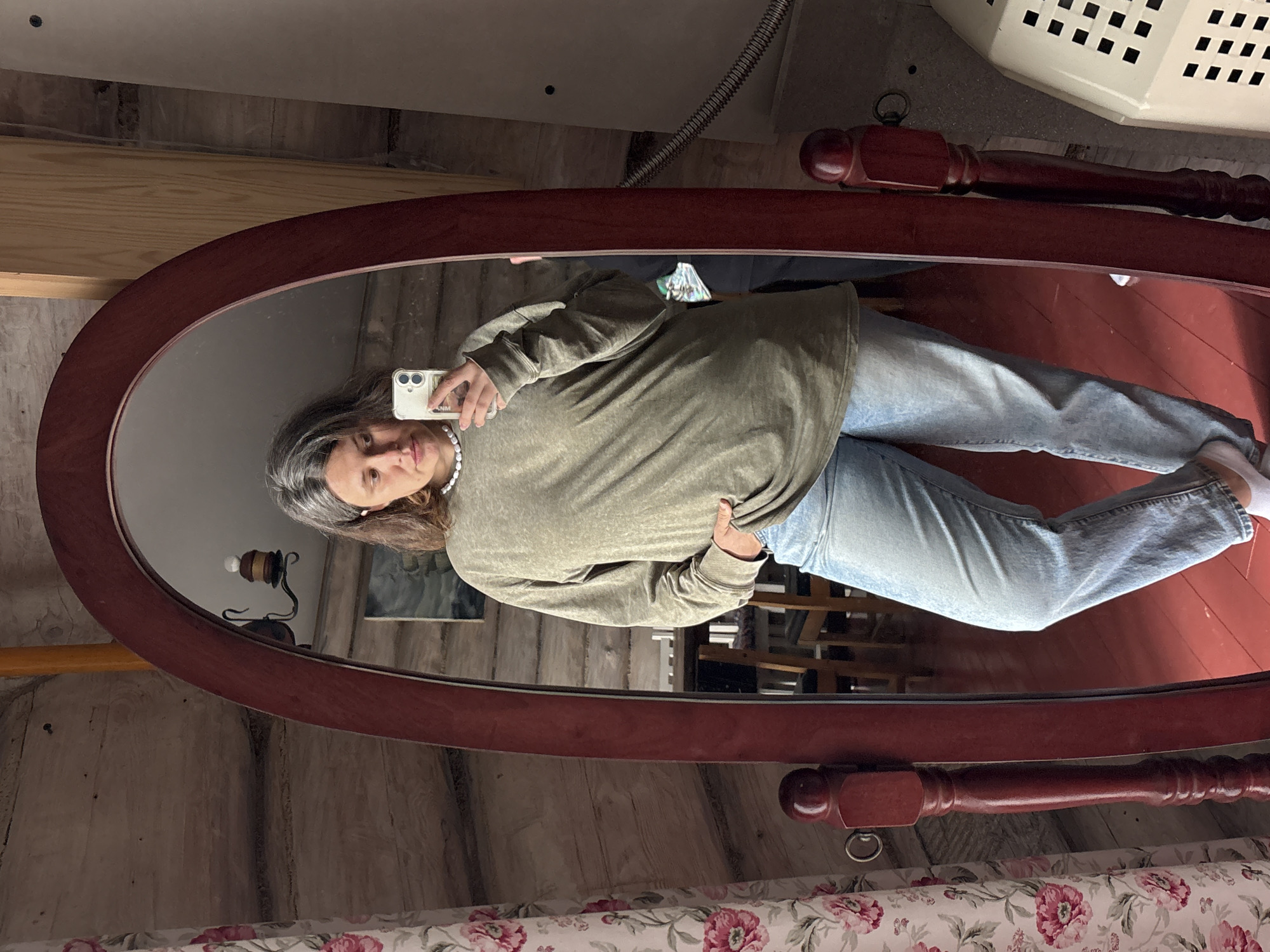 A person is taking a mirror selfie in what appears to be a rustic room. The person is wearing a long-sleeved olive green shirt and light-wash jeans. They are also wearing a pearl necklace and white socks. The mirror is oval-shaped with a dark wood frame. The reflection shows the person holding a white smartphone, capturing the image.
In the background, reflected in the mirror, are wooden walls with visible grain, suggesting a cabin or older building. A vintage-style lamp with a light bulb is mounted on the wall. The floor in the reflection is a reddish-brown, possibly wood or painted. Further in the background, a white laundry basket with a grid pattern is visible, along with a metallic pipe.
The overall impression is casual and personal, with the setting providing a warm, slightly aged ambiance. There is no clear indication of the time of day or weather conditions outside. The person's expression is neutral, focused on taking the selfie. The visible text on the phone case is "ANM". The location is identified as Kostovaty, Russia.