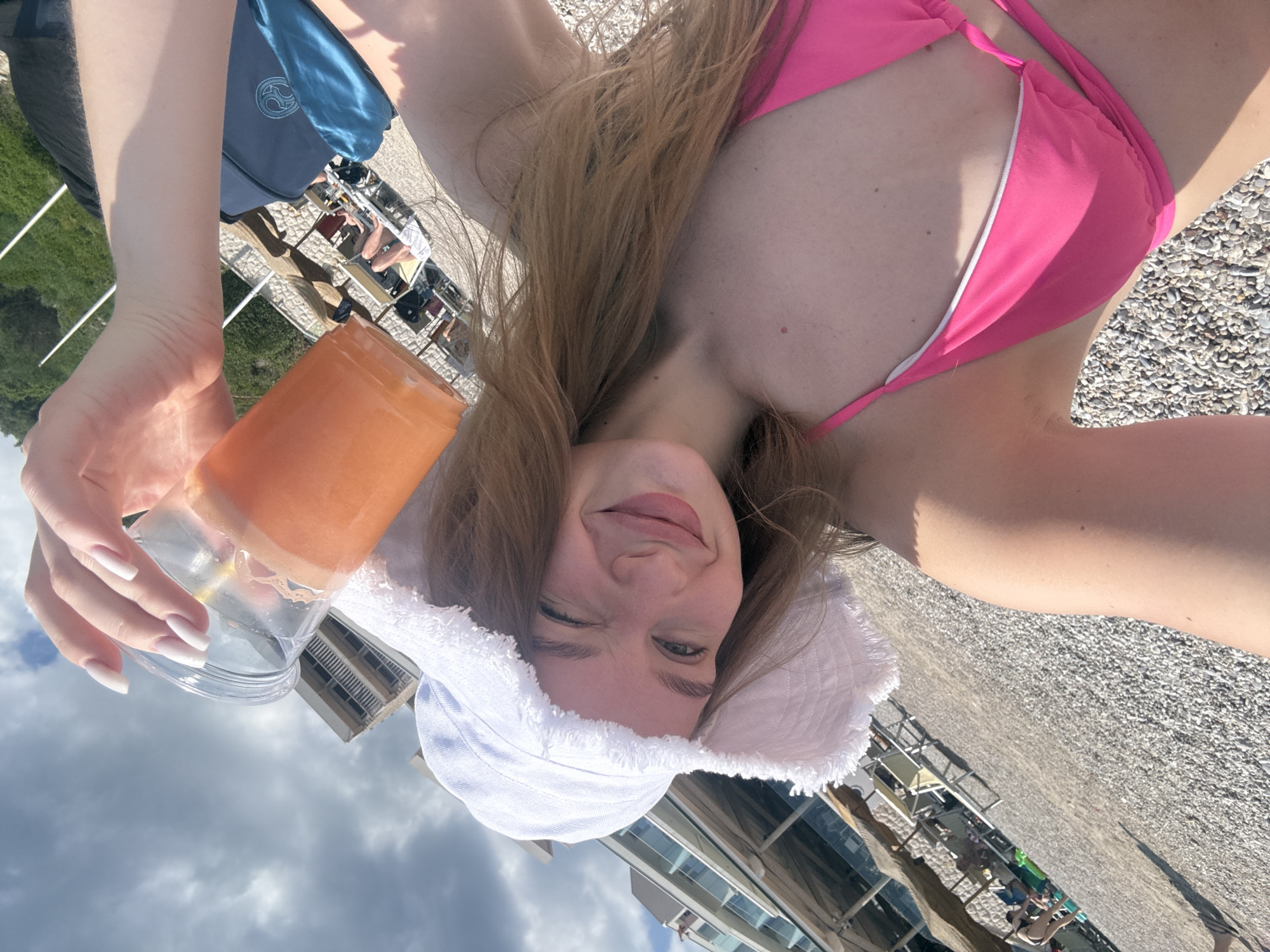 A fair-skinned woman in a pink bikini and white fringed hat is holding a clear plastic cup with an orange liquid in it. She is smiling and winking at the camera. The image is taken on a beach with gray pebbles. Lounge chairs and people are visible in the background. Buildings are present in the background. The sky is cloudy. The image suggests the beach could be near Prijevor, Montenegro.