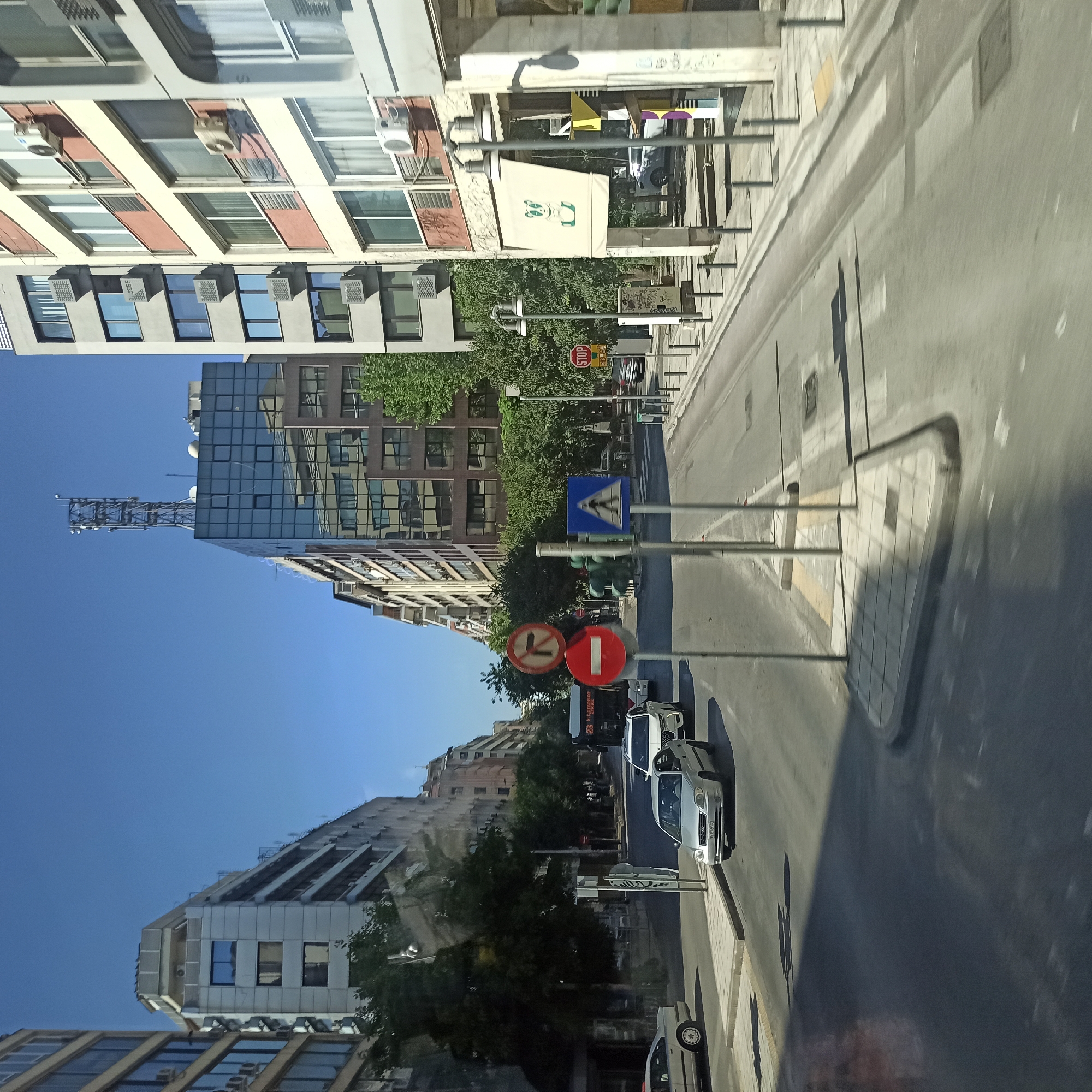 This is a ground-level, eye-level, medium shot of a street scene in Thessaloniki Municipal Unit, Greece, on a clear, sunny day. The perspective is from a vehicle, as indicated by the motion blur and reflection on the lower left. The street is paved with asphalt and has white lane markings. A pedestrian crossing is visible in the foreground.
On the right side of the street, there is a sidewalk with bollards. Buildings line the street on both sides. The buildings are multi-story, constructed with modern architecture, featuring large windows and air conditioning units. One building on the right has a light-colored awning with a cartoon drawing of a panda.
Several vehicles are present on the street. A silver sedan is in the foreground, and a white SUV is behind it. A bus with the number "23" and text that appears to be "Ν. ΣΤΑΘΜΟΣ ΣΙΚΙΕΣ" is also visible. Traffic signs are present, including a "STOP" sign on the right, a pedestrian crossing sign, and two circular signs indicating traffic restrictions. Trees are planted along the sidewalks.
The sky is clear and blue. A tall telecommunications tower is visible on top of a glass-covered building in the distance. The overall impression is of a typical urban street in Greece.