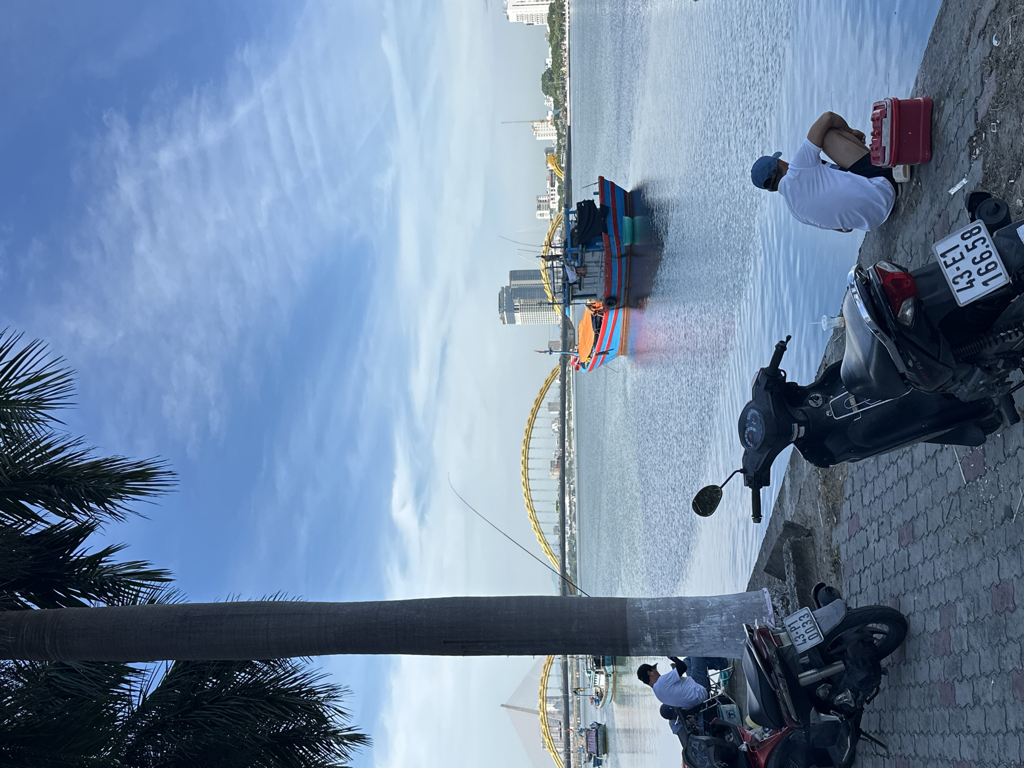 The image depicts a serene waterside scene in Da Nang, Vietnam, likely along the Han River. The foreground features two parked motorcycles, one red and black, the other black, with visible license plates. A person wearing a white t-shirt and a black cap is seated on the pavement, looking at their phone. To the right, another person in a white t-shirt and a blue cap is crouched by the water, fishing with a rod. A red cooler box sits beside them.
In the middle ground, a vibrantly colored fishing boat with blue and red hull and orange canopy is moored on the water. Further in the background, two bridges are visible: a suspension bridge with yellow cables on the left, and the iconic Dragon Bridge, characterized by its yellow arched structure, on the right. The cityscape of Da Nang is apparent on the far bank, with several modern buildings and a construction crane. The sky is a clear blue with scattered white clouds. A large palm tree dominates the left side of the frame, its fronds extending towards the sky. The water is calm, reflecting the sky and the surrounding environment.