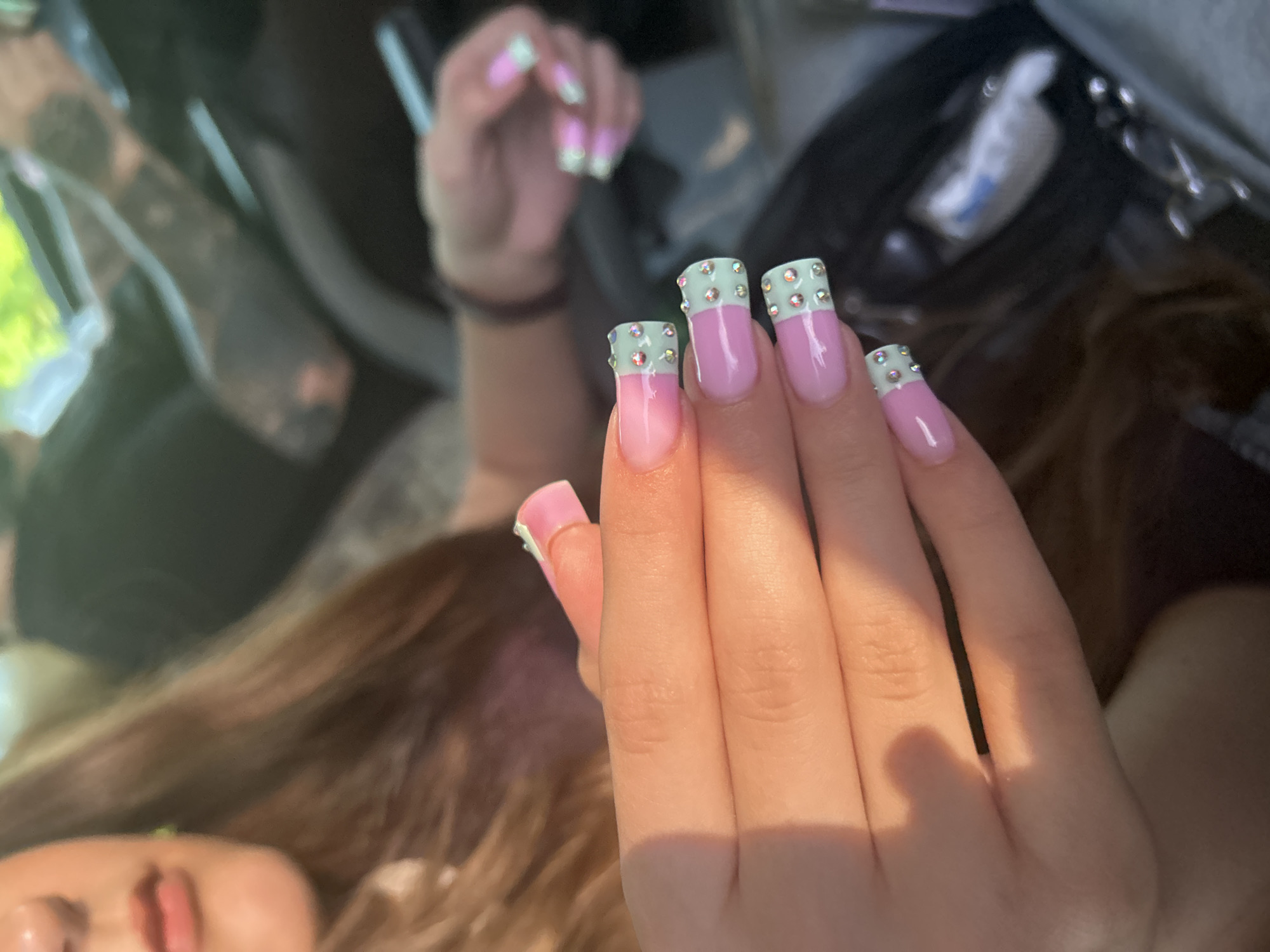 A close-up shot captures a woman's hands adorned with long, pastel pink and mint green French-tip acrylic nails decorated with small rhinestones. The woman, partially visible from the chest up, has brown hair and is seated inside a vehicle. The background shows a blurred view of another person, likely in the driver's seat, and the interior of a car. The lighting suggests it is daytime, possibly late afternoon, casting warm shadows on the woman's hands. There is no discernible information to place this image specifically in Maiaky, Ukraine.