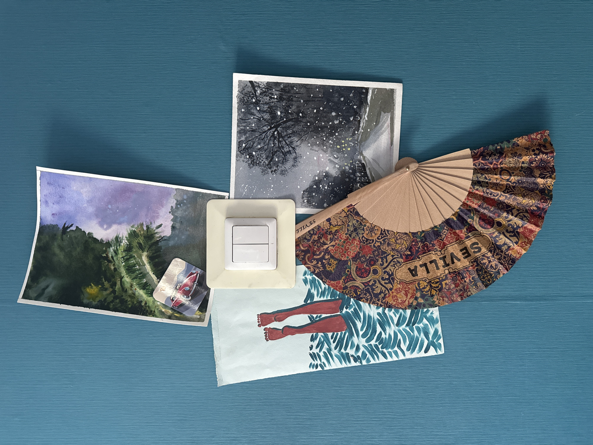 A still-life composition rests on a blue, textured surface, possibly a table or wall. The objects consist of several pieces of artwork, a light switch, and a folding fan. A watercolor painting depicting a landscape with trees and a purple sky is located on the left. A small photograph of a red car is taped to the painting. Adjacent to the painting is a white, rectangular light switch. Above the light switch is a watercolor painting of a snowy, tree-filled landscape. A partially unfolded folding fan with intricate patterns and the word "SEVILLA" printed on it is placed to the right of the snow scene. Finally, a small watercolor painting of bare legs with teal-colored water patterns beneath is placed slightly below the fan and to the right of the light switch. The scene appears to be indoors. There is no evidence that the scene is located in Kazan, Russia.