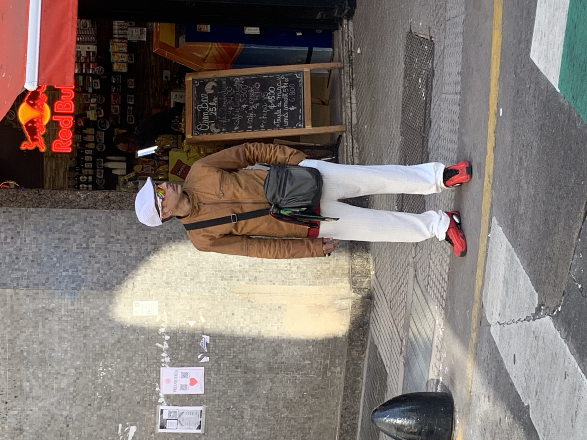 A person stands on a mosaic-tiled sidewalk next to a street in what appears to be Buenos Aires, Argentina, under bright daylight. The individual is a man wearing a brown jacket, white pants, red sneakers, a white cap, and sunglasses. He has a dark grey messenger bag slung across his body.
To his right, there is a small storefront or kiosk, identifiable by shelves stocked with various products and a Red Bull neon sign displaying its logo and text in bright red. In front of the store, a black chalkboard sign advertises "Open Bar" and lists various items and prices in Spanish, such as "Chipa x5," "café," and "trule de miayi," with prices including "$340," "$5300," "$1800," and "$6.800," likely in Argentine Pesos. Part of a "Doritos" logo is also visible inside the store.
The street adjacent to the sidewalk has yellow and white painted lines, indicative of road markings or a pedestrian crossing. A black bollard is visible near the street edge. On the lower part of the wall or ground, two printed signs are visible; one clearly states "PELUQUERÍA" (hairdresser or barber shop). The sunny weather is evident from the distinct shadows cast on the pavement. The overall setting suggests a casual urban street scene.