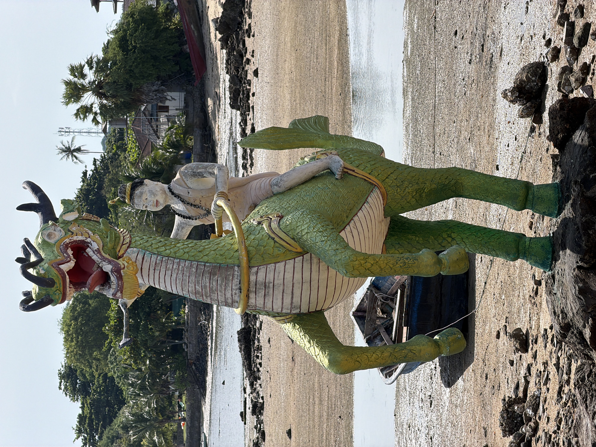 A statue of a green dragon with a rider stands on a sandy beach in Baan Plai Laem, Thailand. The dragon is depicted with its mouth open, a long, red tongue, and antlers on its head. The rider is a small, light-skinned figure wearing a brown garment and a necklace. The dragon is painted in green and white, with yellow accents on its saddle and mane.
The beach is covered in sand and small rocks. A small, blue boat is overturned on the sand behind the dragon. In the background, there are trees, palm trees, and some buildings, including a cell tower. The sea is visible in the distance. The sky is clear and blue.