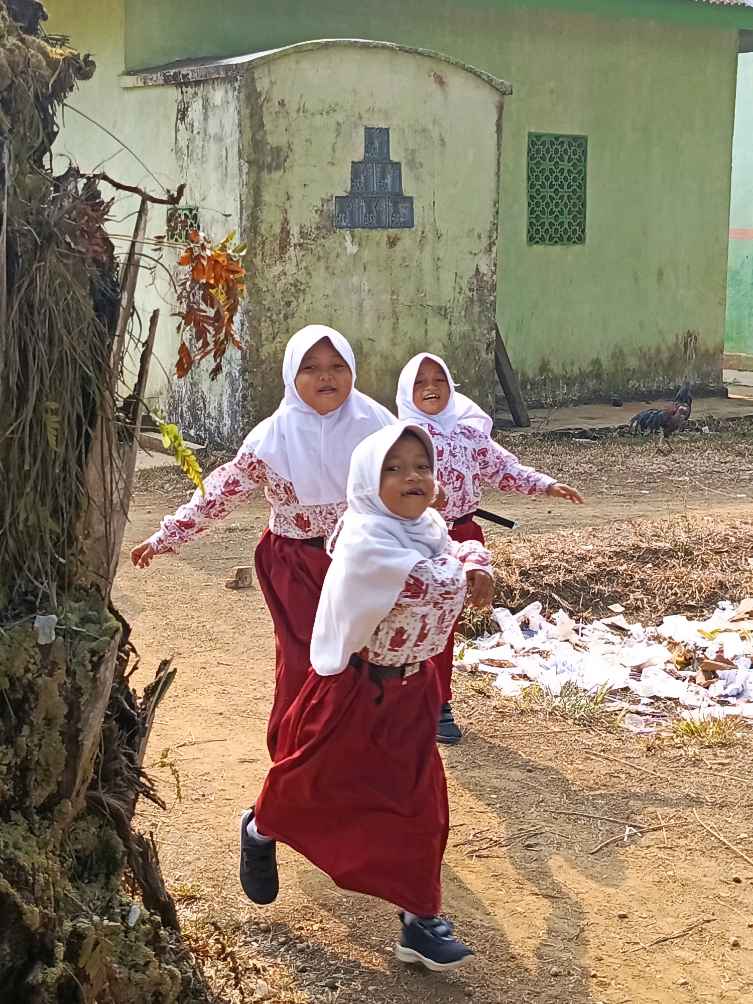 Three young girls wearing white hijabs and maroon skirts with red and white patterned tops are captured in motion outdoors. The girl in the foreground is facing forward with her mouth open, seemingly mid-sentence, with her right arm extended and her left arm bent. The middle girl is looking to her right and smiling, with both arms extended outward. The third girl, partially visible in the background, is also smiling and looking to her left. They appear to be in a schoolyard or open area with a dirt ground. In the background, a green building with a weathered facade and a green decorative window is visible. To the right of the building, a rooster is standing on the dry grass. A large, moss-covered tree trunk is prominent on the left side of the frame. The scene is illuminated by natural sunlight, casting shadows on the ground. The presence of scattered white plastic debris in the foreground suggests a potentially unkempt environment. This image was taken in Megang Sakti IV, Indonesia.