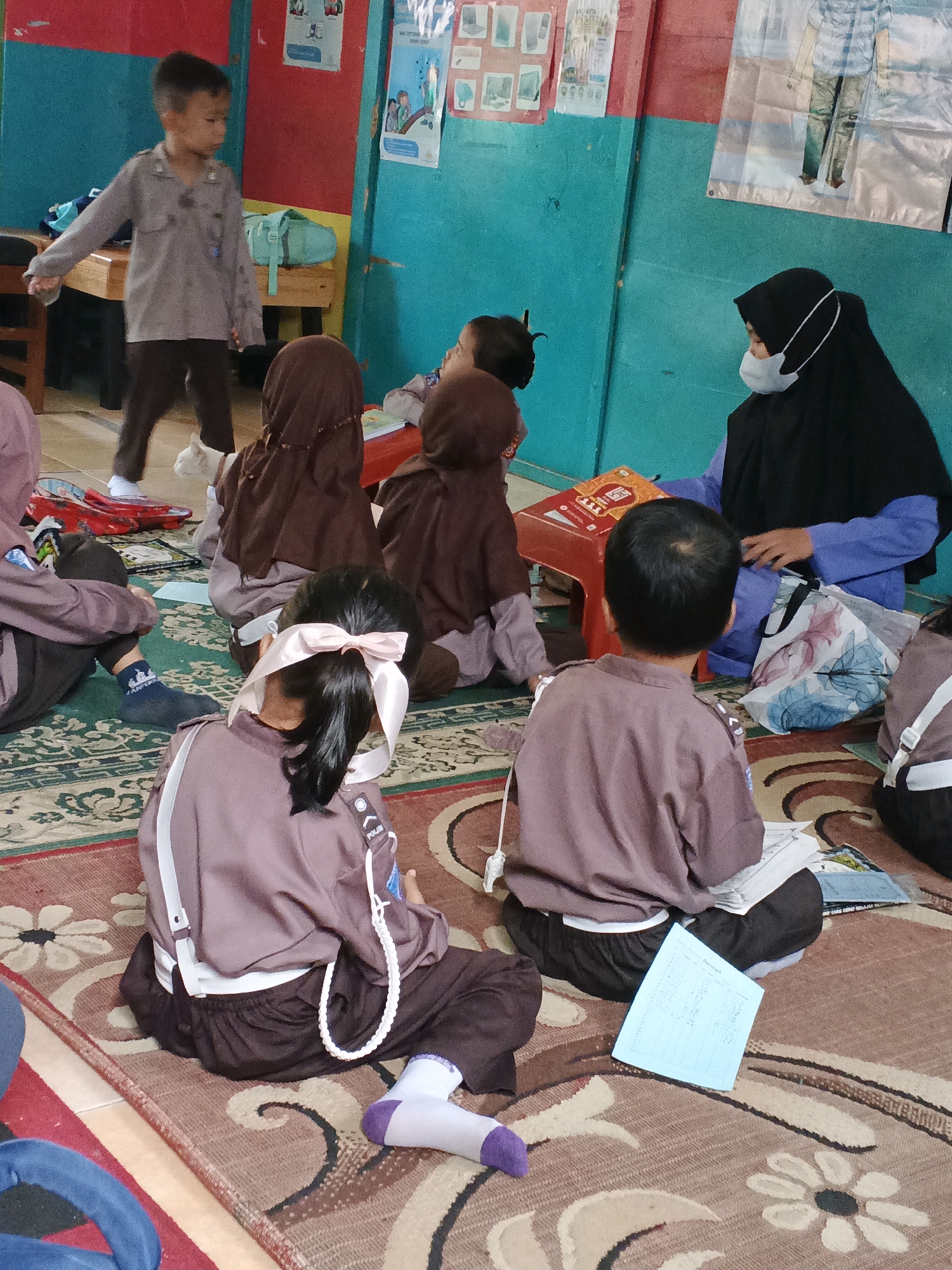 The image depicts a classroom scene at a school in Warungkiara, Indonesia. Several children, dressed in uniforms with brown shirts and dark pants or skirts, are seated on the floor on a patterned rug. Some of the children are wearing headscarves. One young boy in a grey uniform stands to the left, looking towards the seated children. A woman wearing a black hijab and a blue top is seated on the right, interacting with a child. On a red plastic stool in front of her is a stack of books. Various educational materials, including posters on the wall and papers on the floor, are visible in the background. The walls are painted in contrasting colors of blue and red.