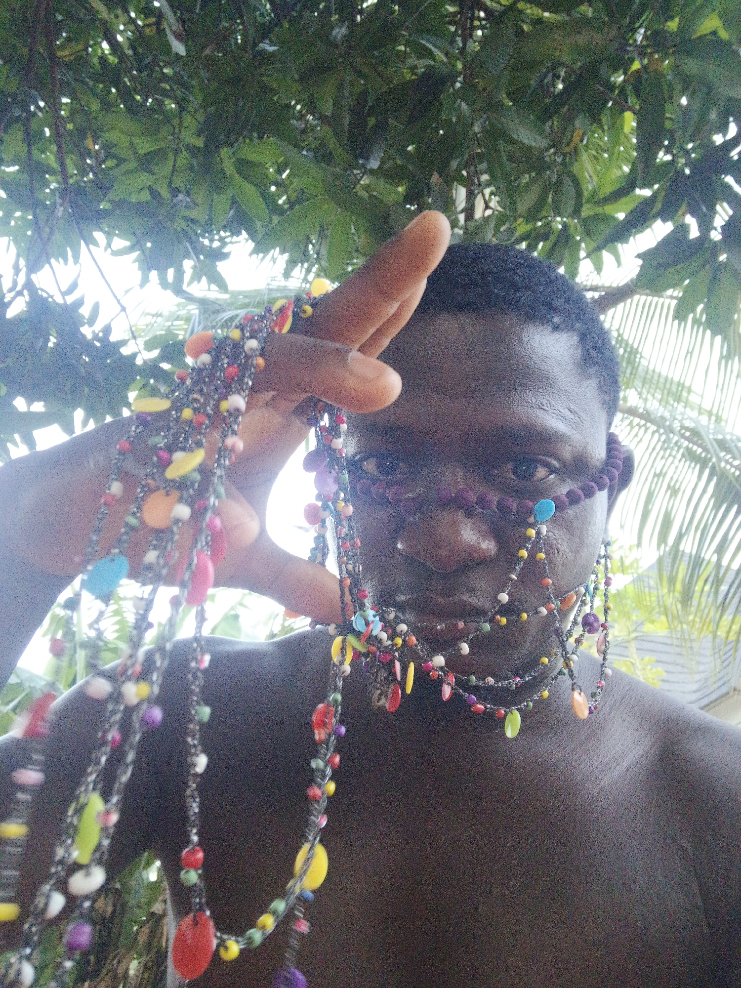 A medium outdoor shot shows a shirtless, dark-skinned person, likely male, holding a string of colorful beads in his left hand and wearing purple beads around the bridge of the nose. A single blue bead is attached to the nasal string of beads. More colorful beaded strands hang from the nose string of beads. The person appears to be outside, with green leaves and some palm trees visible in the background. The location is suggested to be Akwanga, Nigeria.