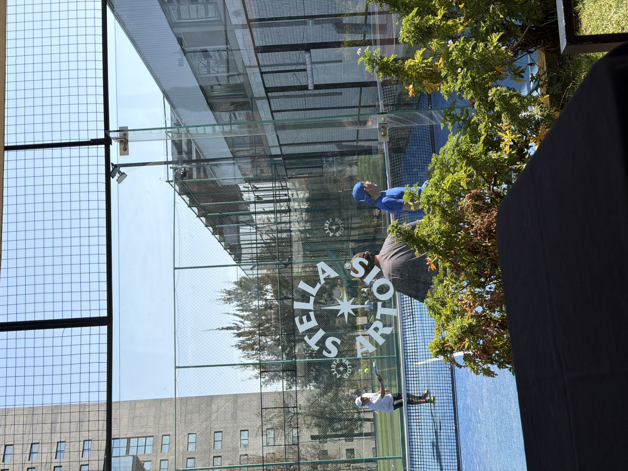 A vertical outdoor medium shot captures a paddle tennis match through a glass pane displaying "Stella Artois" branding. In the background, two players are visible: one wearing a blue cap and another with a brown shirt. In the foreground, a player in a white shirt is in motion, striking a ball. The backdrop includes urban architecture, with buildings visible through the glass and netting surrounding the court. There's also greenery, with a tree branch and a large dark shape (possibly a person or object) partially obscuring the view. The scene is set in the city of Puebla, Mexico.