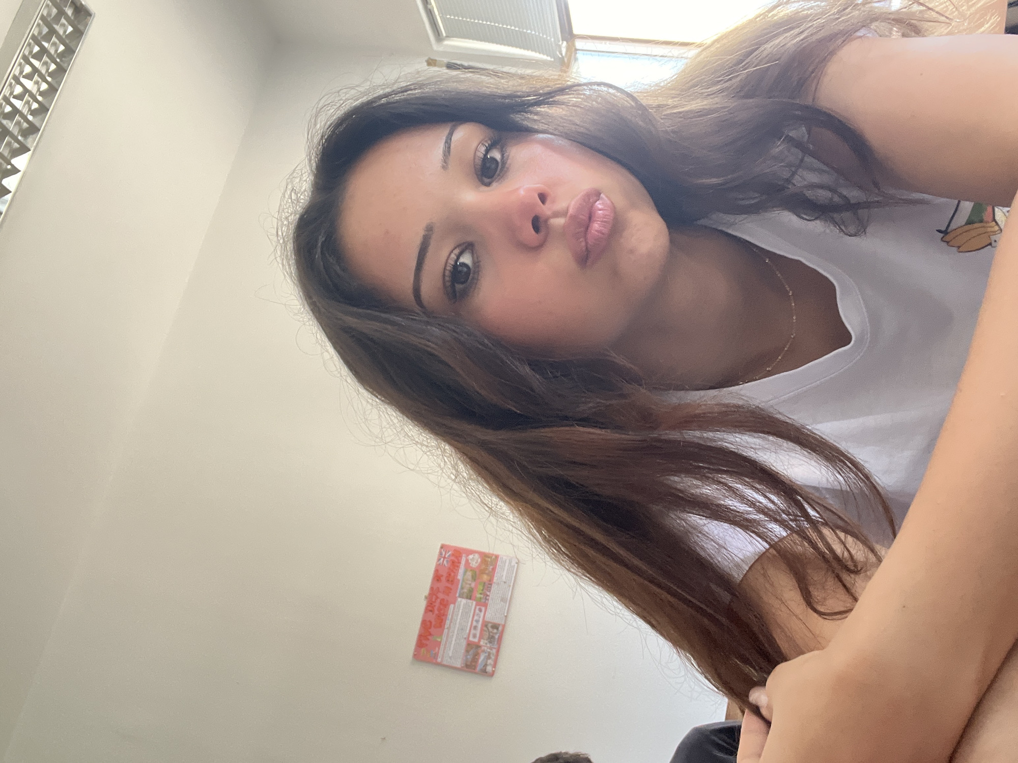 A young woman with long, dark brown hair and light brown skin is captured in a close-up selfie taken indoors. She is wearing a white t-shirt and a delicate gold necklace. Her lips are puckered, and she is looking directly at the camera with a slightly playful expression. Her left arm is resting across her chest, with her hand gently touching her hair. The background is a plain, off-white wall, with a fluorescent light fixture visible on the ceiling. To the left of the woman, a small, colorful poster with text and images is affixed to the wall. The overall lighting suggests an indoor setting, possibly a classroom or office, with natural light filtering in from a window on the right side of the frame. There is no visual information within the image that would allow for the identification of the specific location within Zlatograd, Bulgaria.