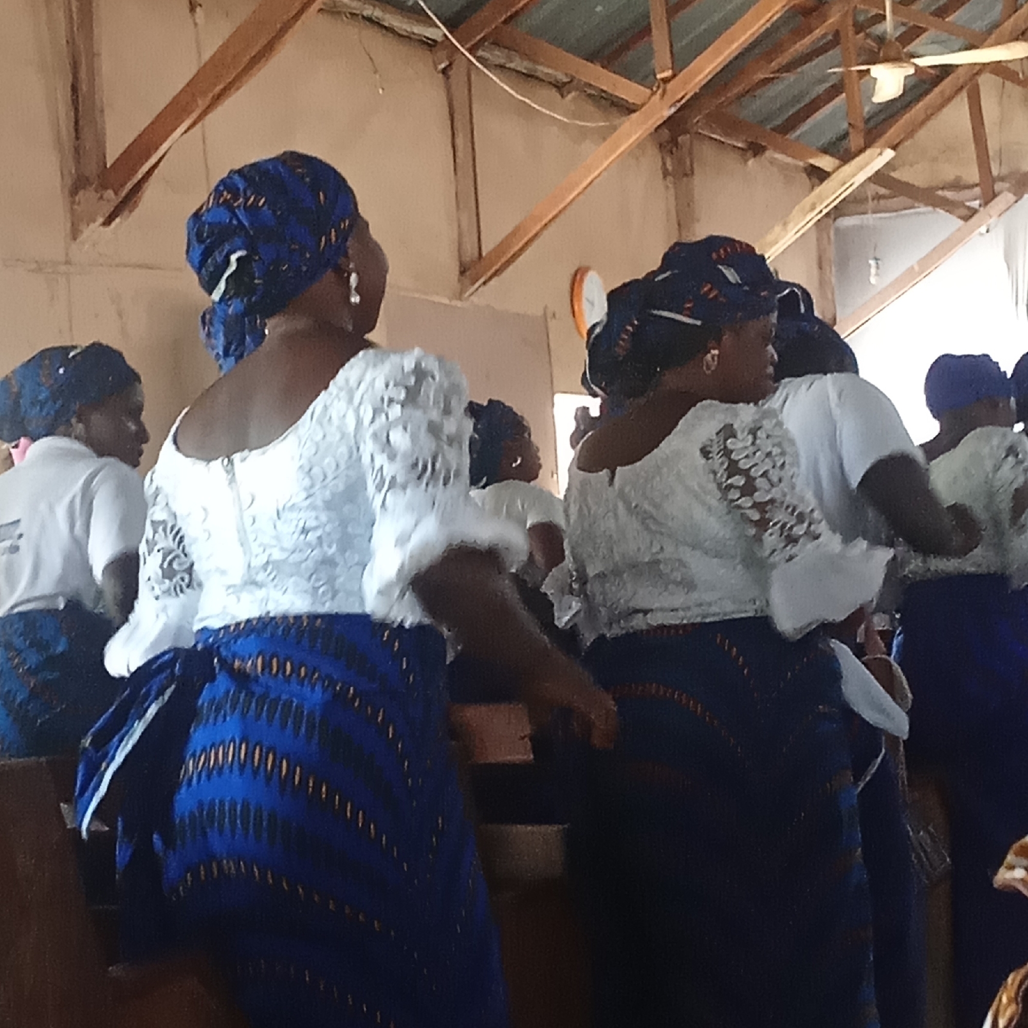 In an indoor space, possibly a church, a group of women are seen from the back, dressed in what appears to be a uniform of white lace tops and blue patterned skirts. They also wear matching blue headwraps. The room has a simple structure with wooden beams visible at the top. There is no clear indication of the location or city.