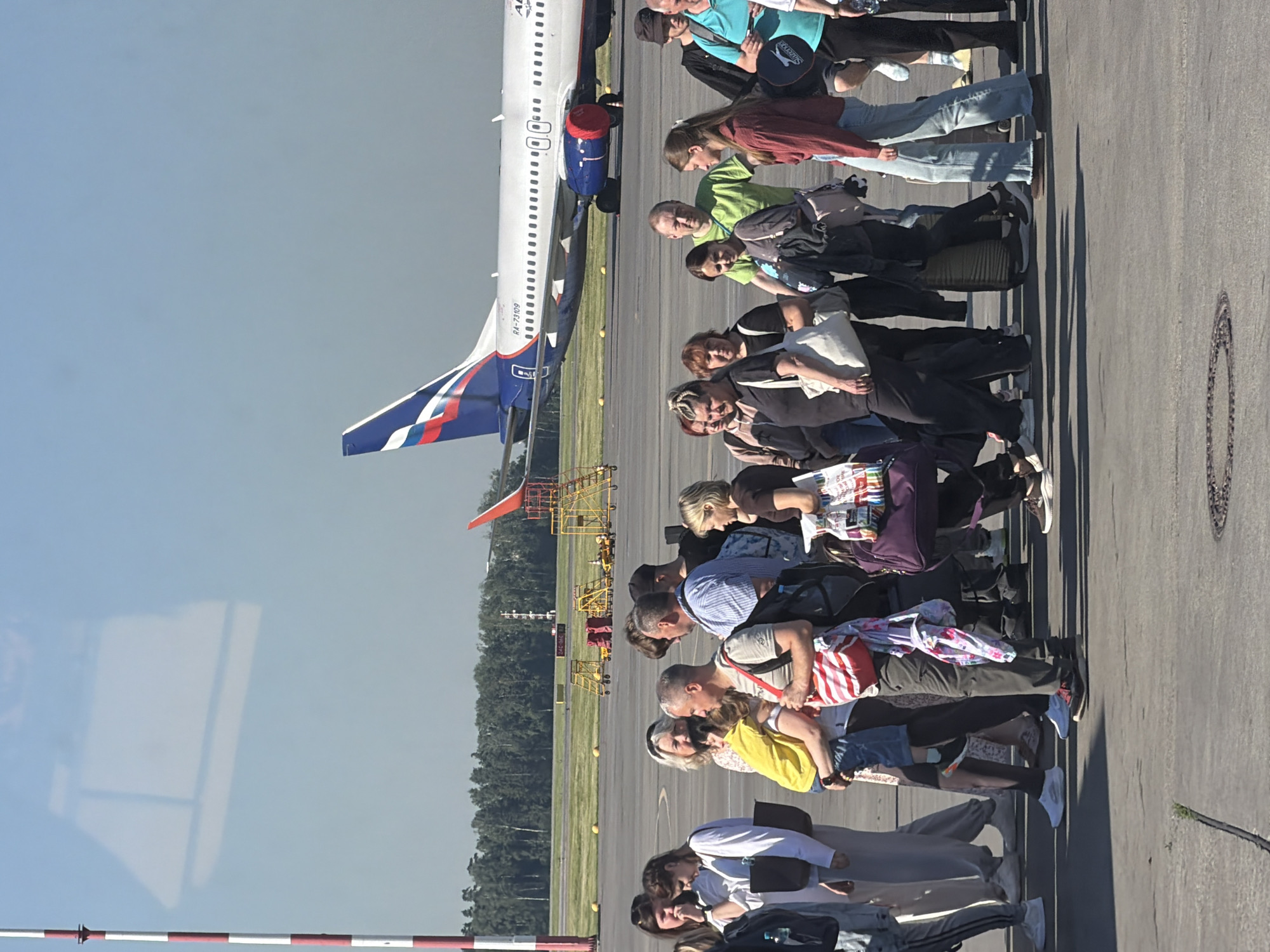 In Khimki, Russia, on a bright, sunny day at an airport tarmac, a group of people are waiting in line, presumably to board a plane. An Aeroflot airplane with the registration RA-73109 is visible in the background, parked on the tarmac. The people, dressed in casual attire, are carrying various bags and luggage. Some are conversing, while others appear to be looking ahead. The airport infrastructure, including service vehicles and a red and white striped pole, are also present in the scene. The ground is paved with asphalt, and the sky is clear blue.