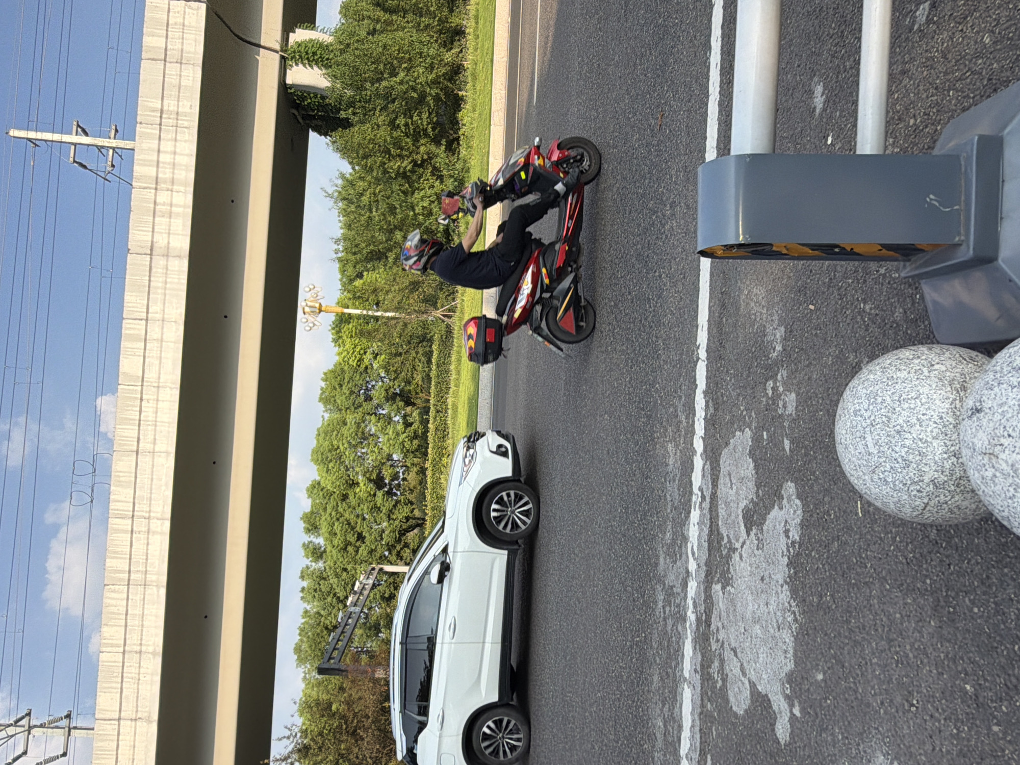 In Jiangjin, China, a white SUV is captured in motion on an asphalt road, with a rider on a red and black electric scooter also moving forward. The scooter has a black storage box on the back. The scene is framed by a large overpass overhead, supported by concrete pillars partially obscured by green foliage. Electrical power lines are visible above the overpass against a clear blue sky with scattered clouds. The road surface shows white lane markings. In the foreground, a grey metal barrier with reflective yellow and black stripes is positioned near two large, speckled grey spherical bollards.