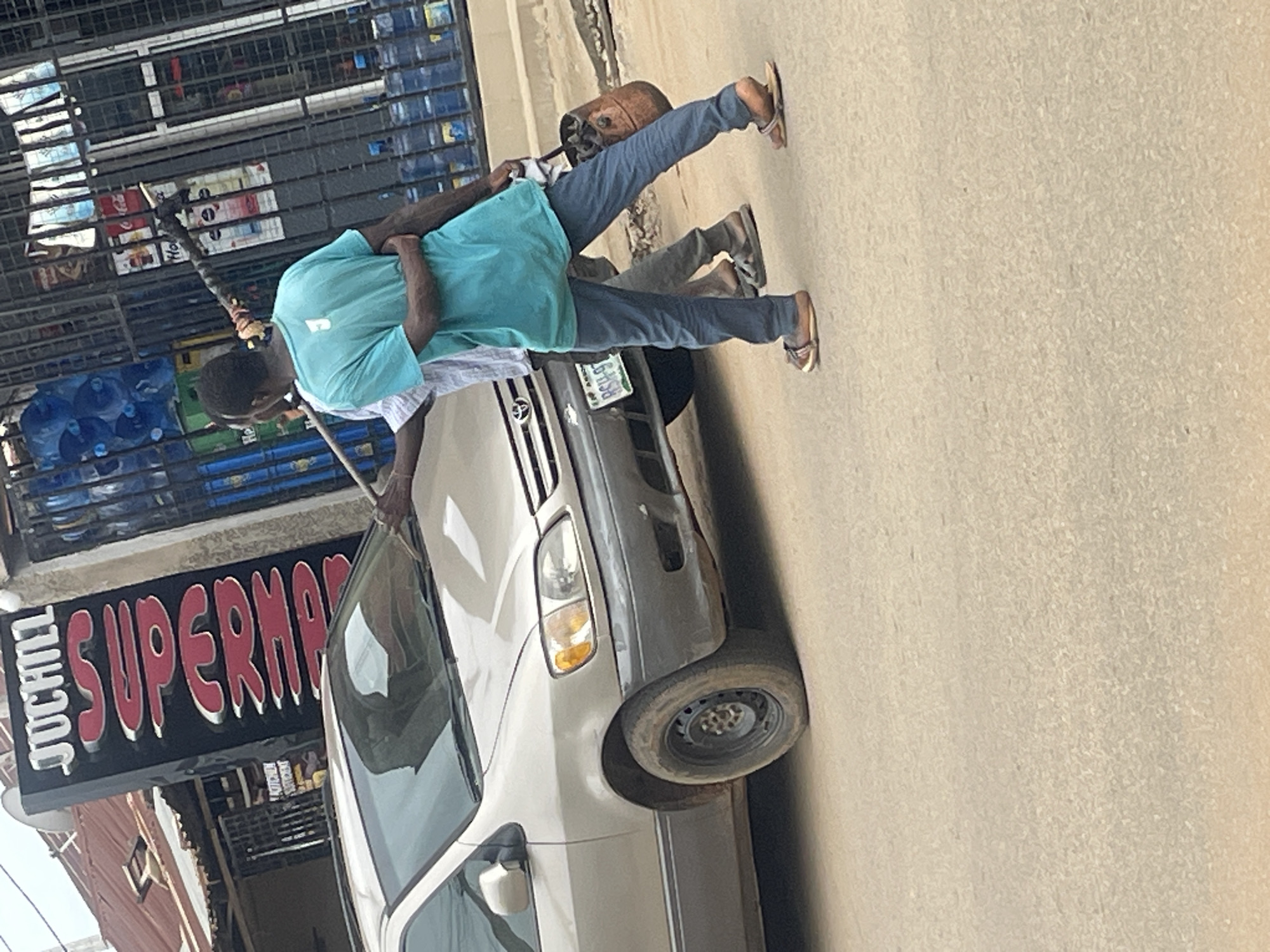 In a street scene in Abuja, Nigeria, a light-colored minivan is parked on a dusty road. A man wearing a teal t-shirt with a white number "J" on the back and blue jeans is seen from behind, with his left arm around another person who is partially obscured. He is holding a long pole with a brown bag attached to one end. The second person, wearing a light-colored shirt and grey pants, is also facing away and has their left arm around the first man's shoulders. They are both wearing sandals. Behind them, a store with a large black sign featuring red lettering that reads "JUCHILL SUPERMARKET" is visible. The store is secured with a metal gate, behind which stacks of bottled water are arranged. The overall environment appears to be an urban street with visible signs of commerce and everyday life.