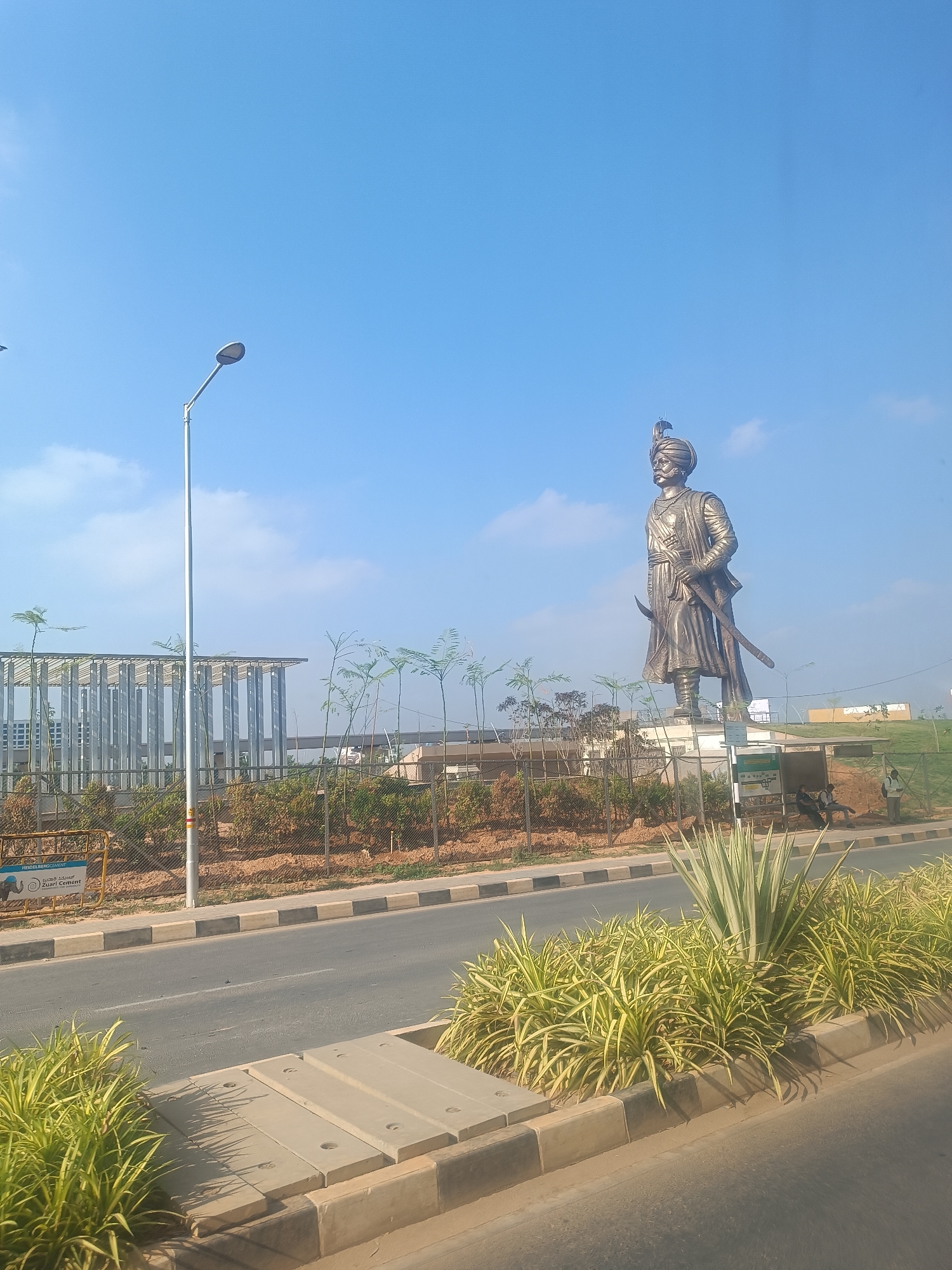 The image depicts a bright, sunny daytime scene in Yartiganahalli, India, showcasing a prominent bronze-colored statue of a historical figure. The large statue stands tall, portraying a man adorned in traditional attire, including a turban, a long tunic, and holding a sword. He features a mustache and beard, facing towards the right of the frame.
In the foreground, a two-lane road runs horizontally across the image, separated by a well-maintained central median. This median is densely planted with decorative green and yellow foliage, including spiky plants, and incorporates concrete drain covers. A modern street light, with a sleek, curved head, stands prominently on the left side of the road.
Behind the median and a chain-link fence, there's a landscaped area with small, young trees and bushes. To the far left, a contemporary building with an open, skeletal metallic structure is partially visible. Near the base of the statue, on the right, an open-sided shelter, likely a bus stop, is present. A couple of people are visible here, one sitting and another standing, suggesting a brief waiting or resting activity. Beyond the shelter, a green, grassy slope is visible.
The sky is a clear and vibrant blue with only a few wispy white clouds, indicating fair weather and strong sunlight that casts distinct shadows from the statue and other objects.
Visible text on a yellow barrier or board on the left side of the road clearly reads "HEIDELBERGCEMENT" and "Zuari Cement," accompanied by an elephant logo. Further text on a sign near the bus stop shelter is present but too small to be legible. The scene suggests a developed and well-maintained public area.