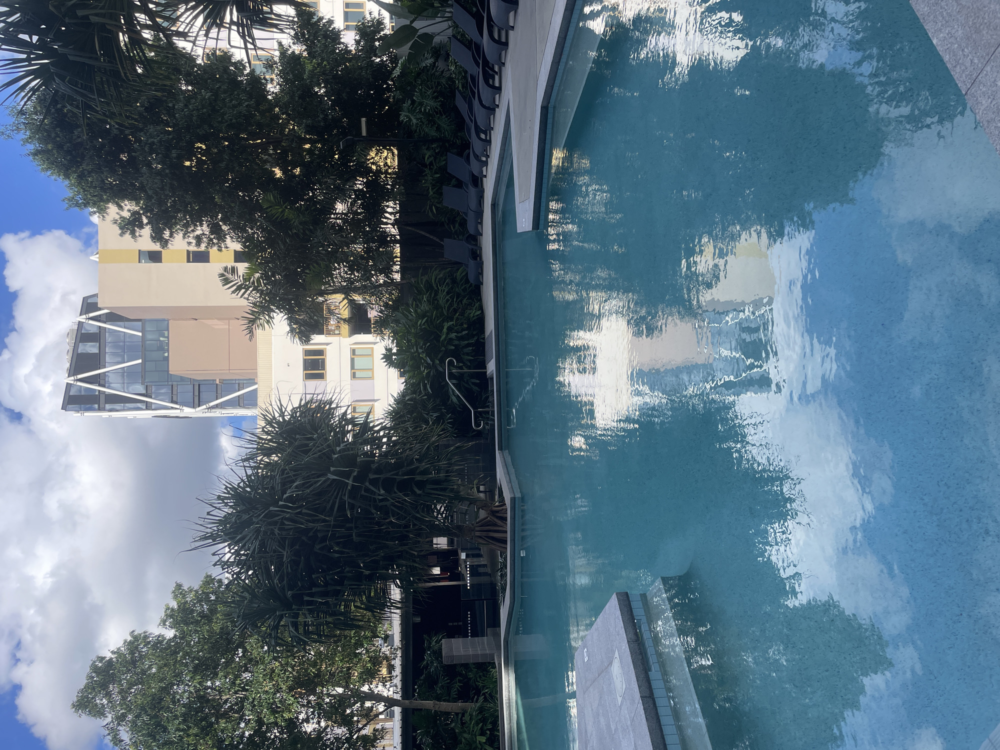 An outdoor swimming pool is shown in the foreground. The pool is filled with bright blue water and reflects the sky and surrounding environment. Around the pool are lounge chairs and lush green plants. In the background, there are several modern buildings. The scene appears to be in an urban setting, likely a hotel or residential complex. This image was taken in Brisbane, Australia.