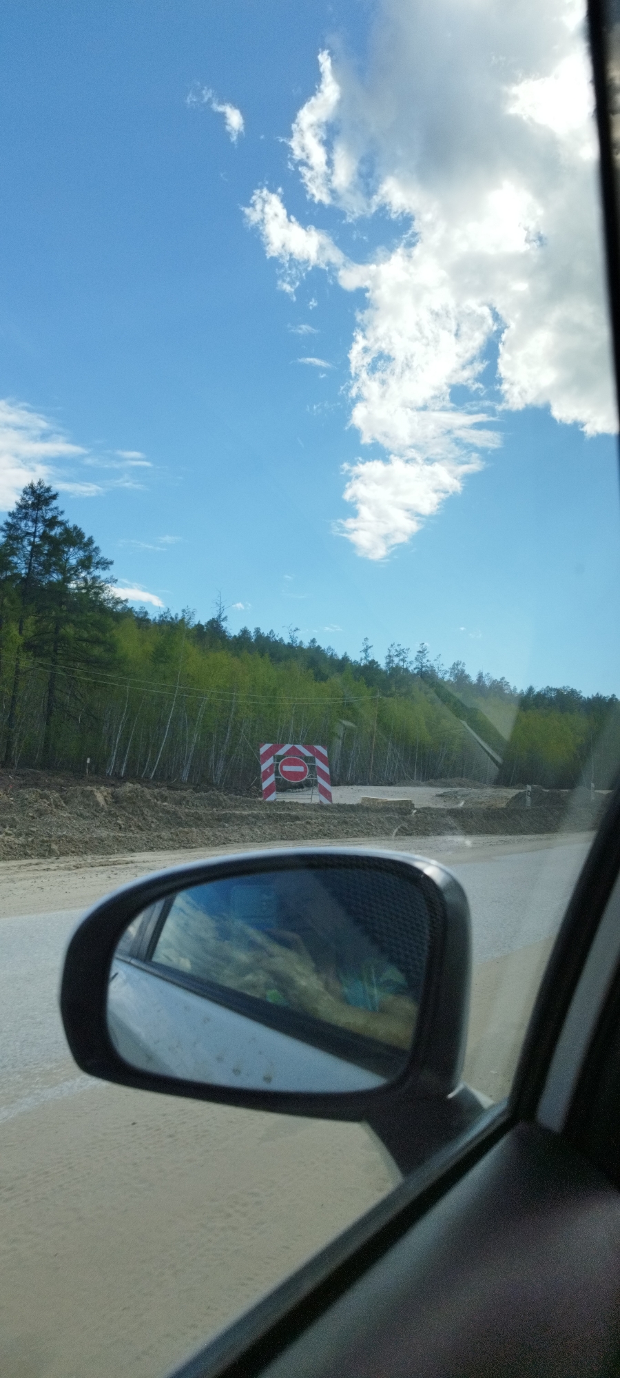 The image, likely taken from a moving vehicle, shows a road in Tabaga, Russia, with a "no entry" road sign placed on a dirt bank, suggesting road construction or closure. The sky is blue with scattered clouds, and a forest line is visible in the background. The car's side mirror and interior are partially visible, framing the outdoor scene.